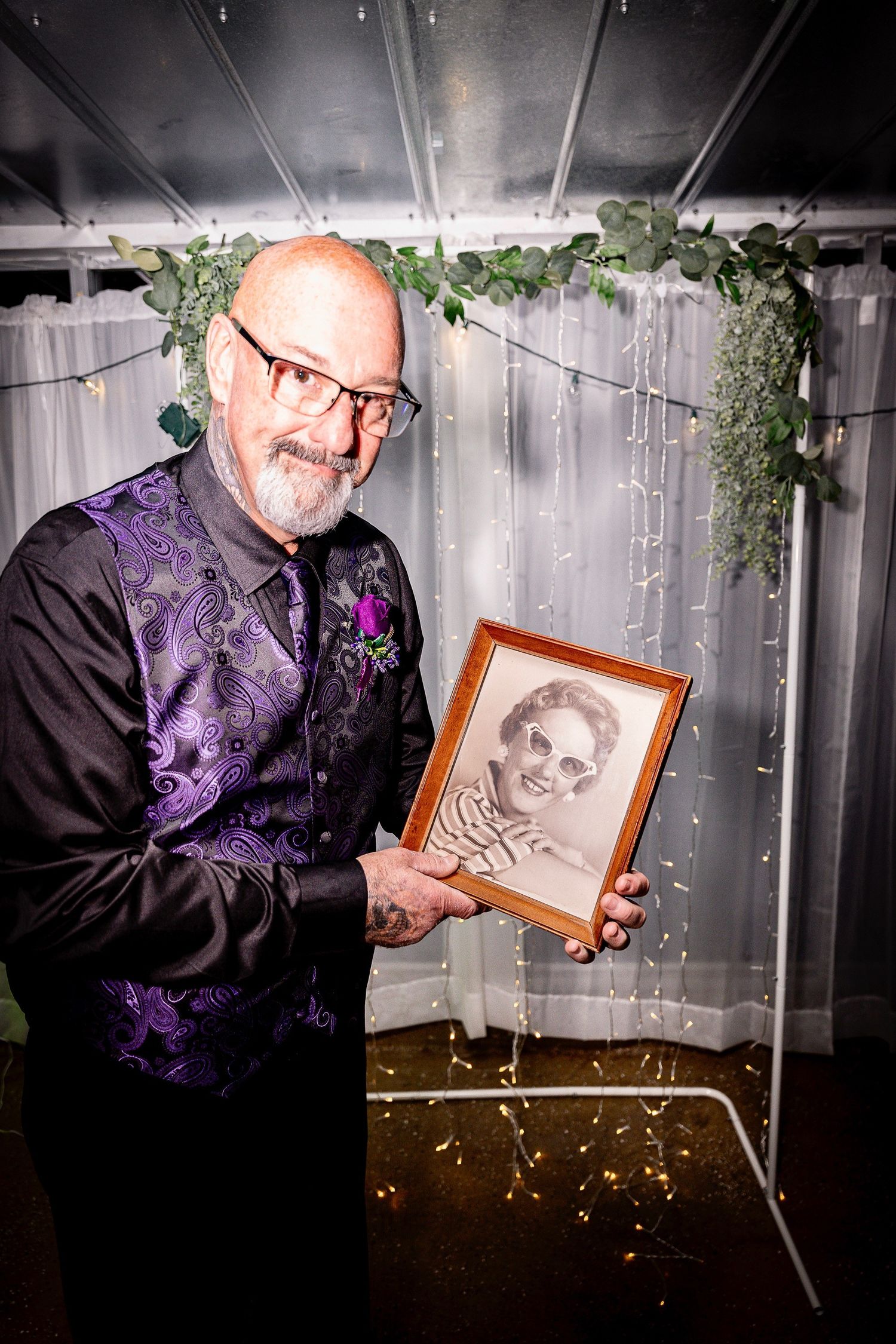 Person in formal attire holding a framed photograph against a silver backdrop with twinkling lights.