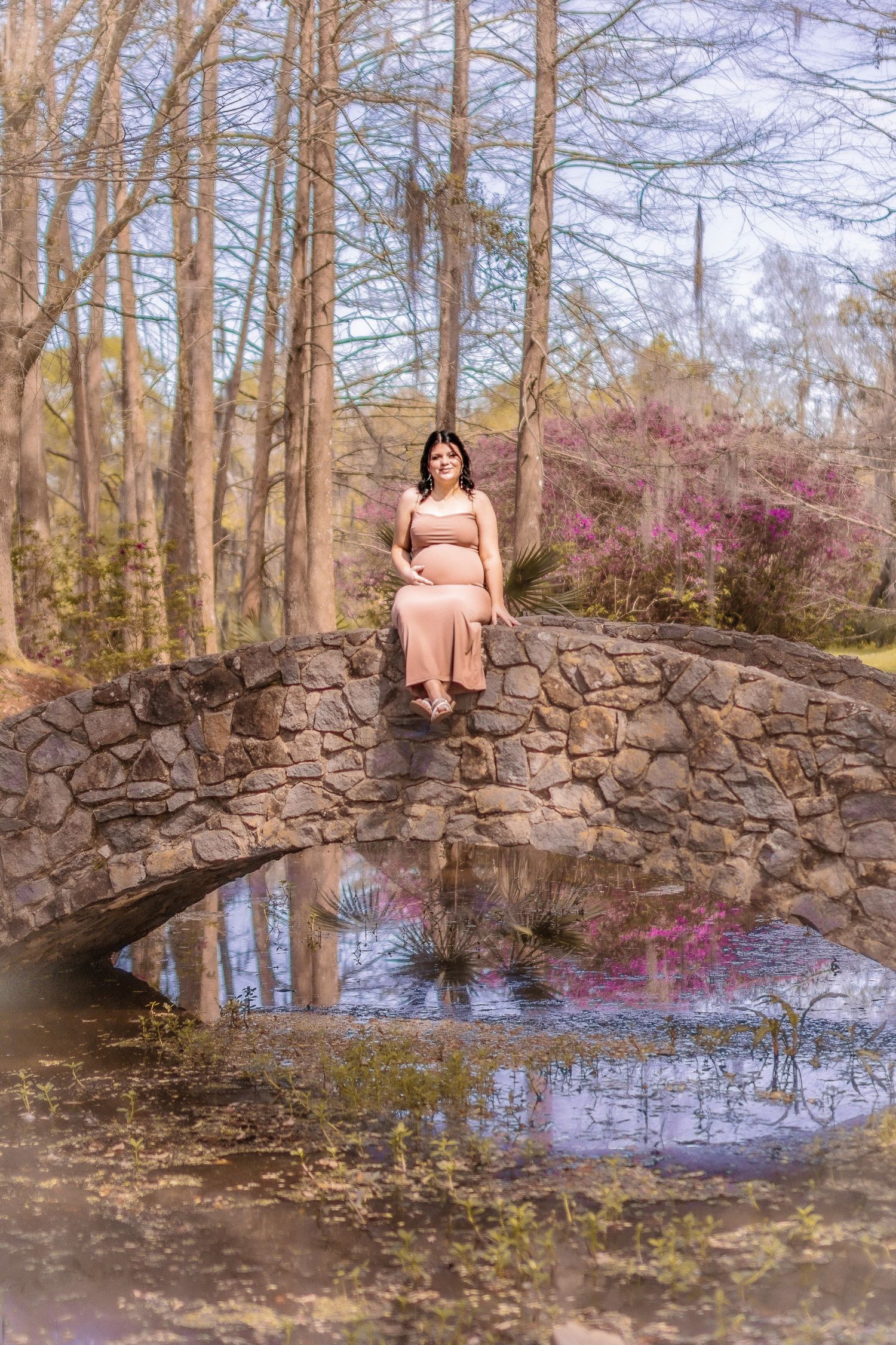 Person in pink dress poses on stone bridge over pond with blooming spring trees in background.
