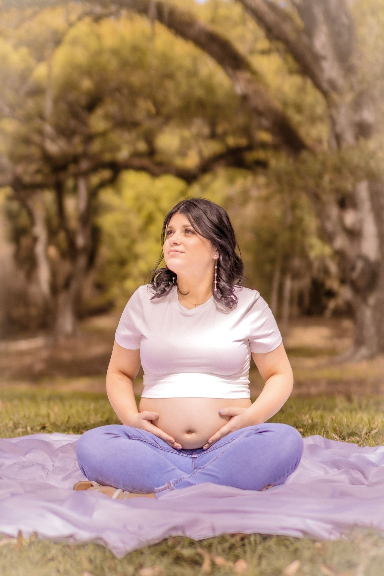 A person in a white t-shirt and purple pants sits cross-legged on a purple blanket in a park setting with autumn trees.