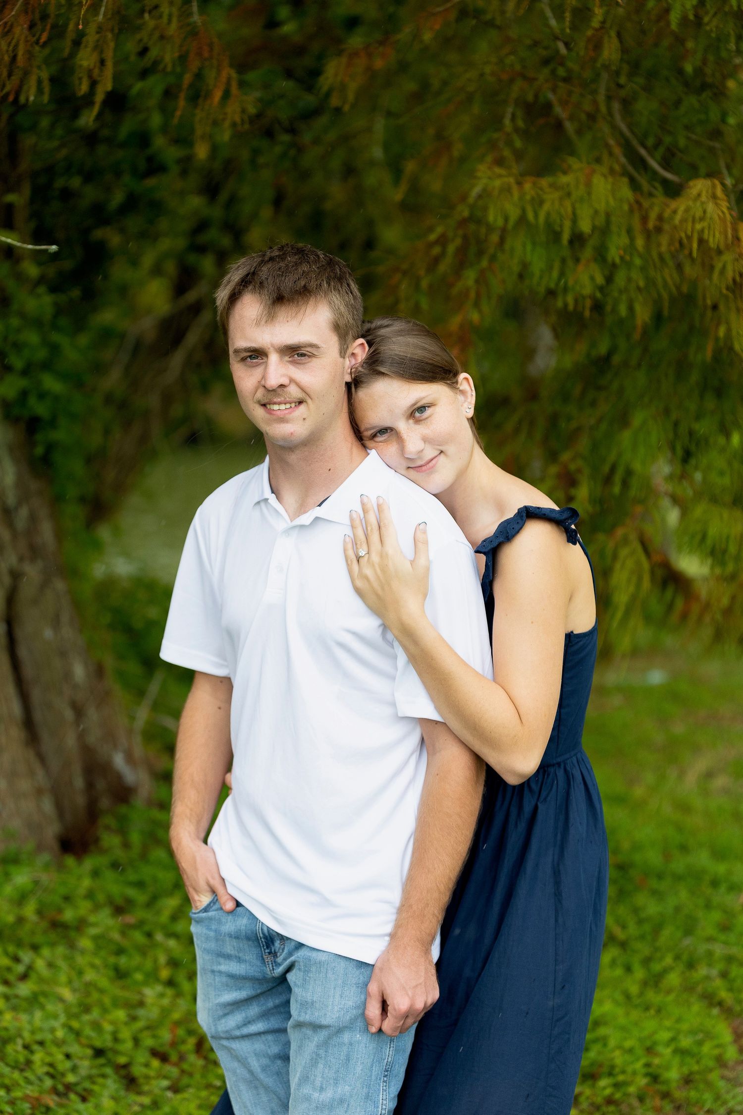 A couple embraces outdoors in casual attire against a natural green background.