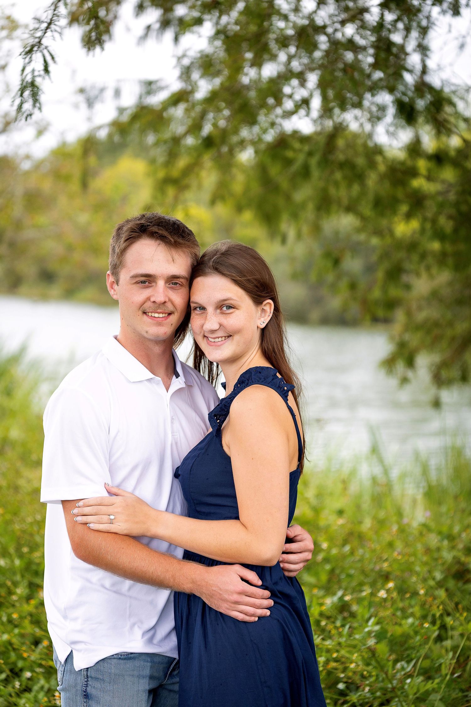 Romantic outdoor couple photos by lake with overhanging trees in background.