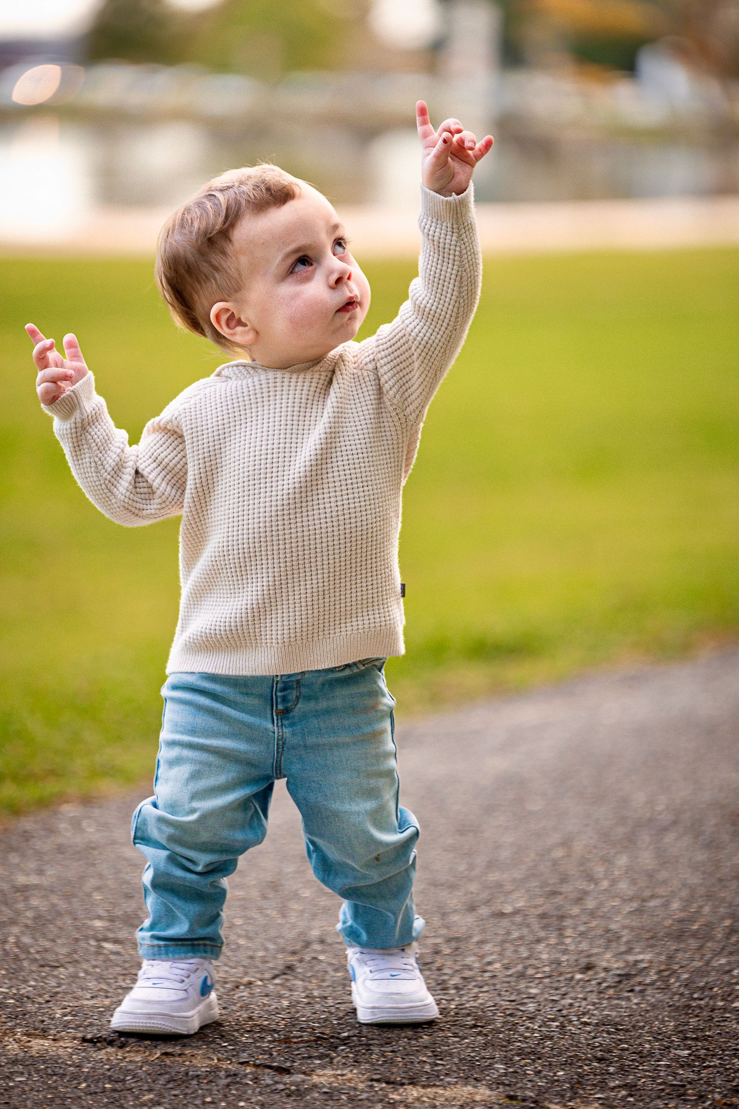 Toddler in cream sweater and jeans pointing upward while standing on path.