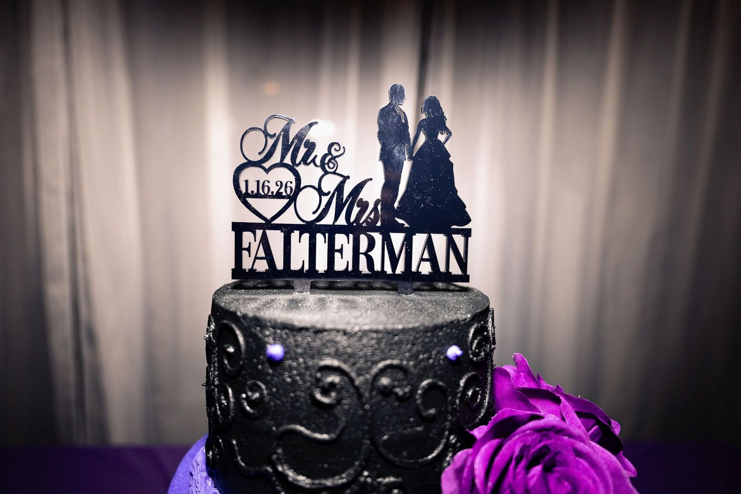 Wedding cake with black frosting details and purple accents on white backdrop.