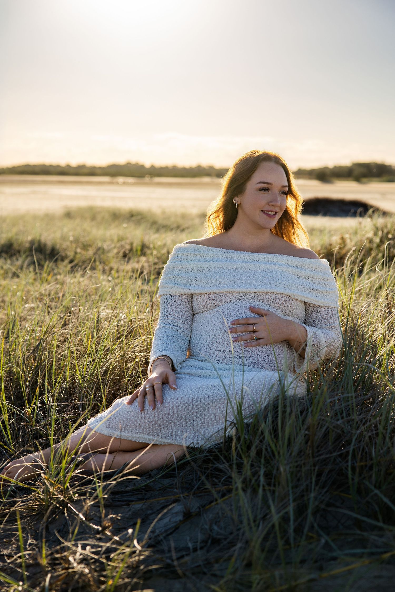 Pregnant woman in white knit off-shoulder dress sitting in golden hour sunlit field, cradling baby bump.