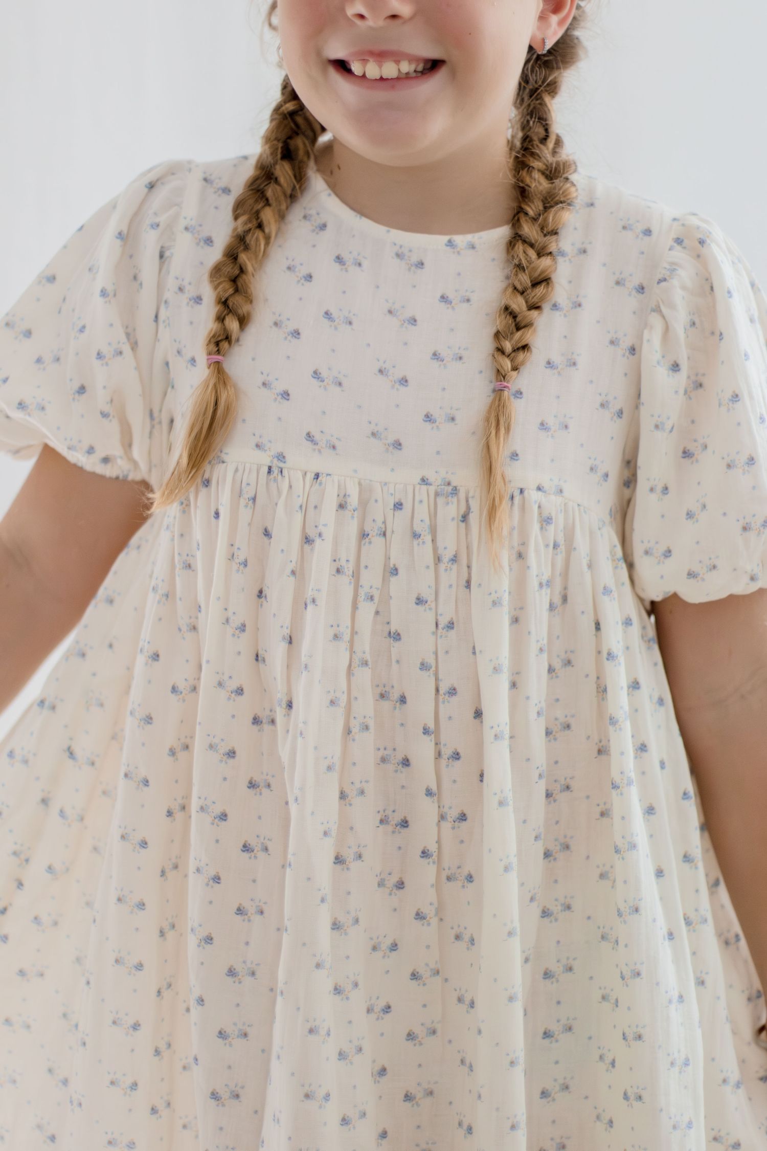 Smiling young girl with braids wearing cream floral print puff sleeve dress against white background.