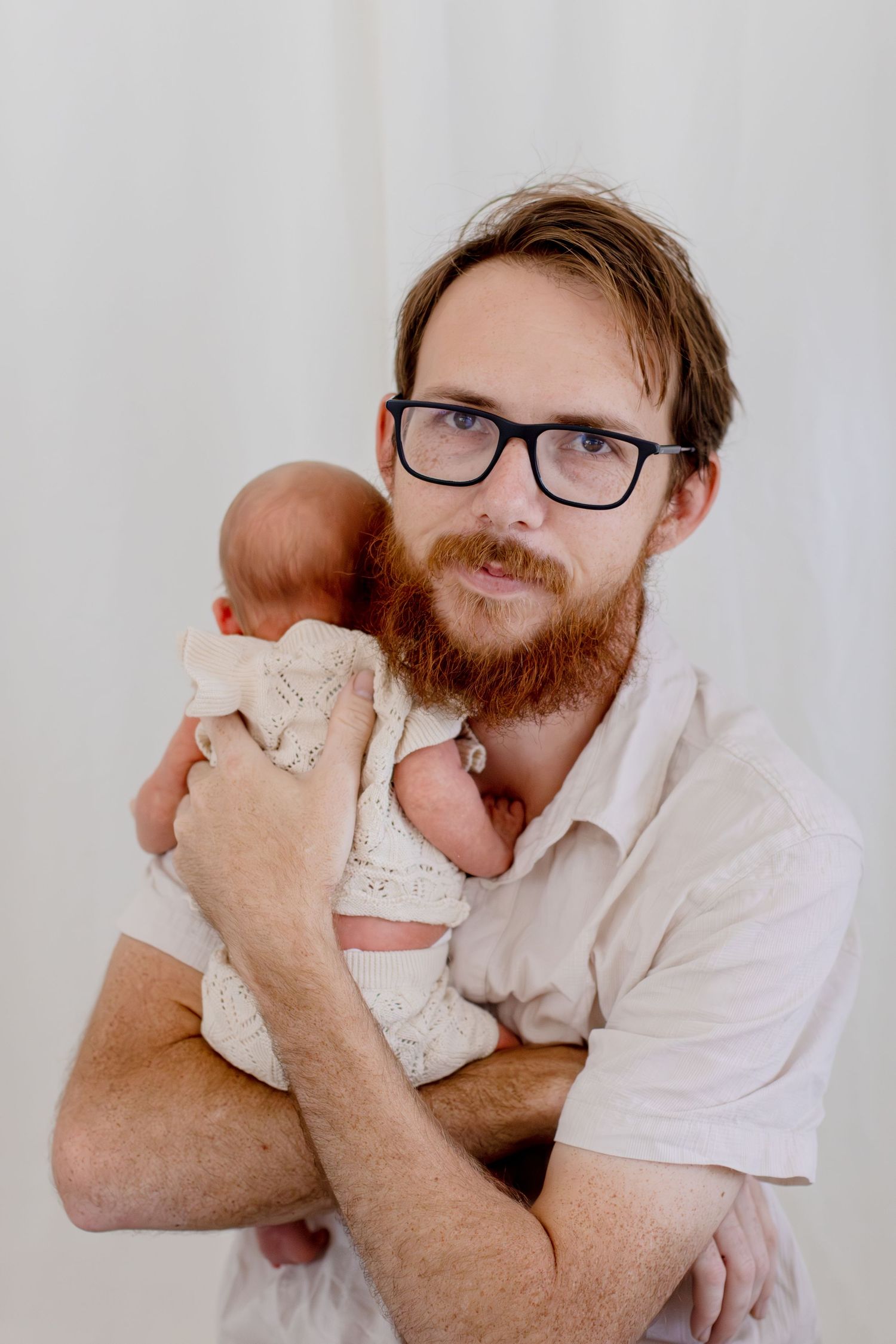 Bearded father with glasses tenderly holds sleeping newborn baby in cream knit wrap.
