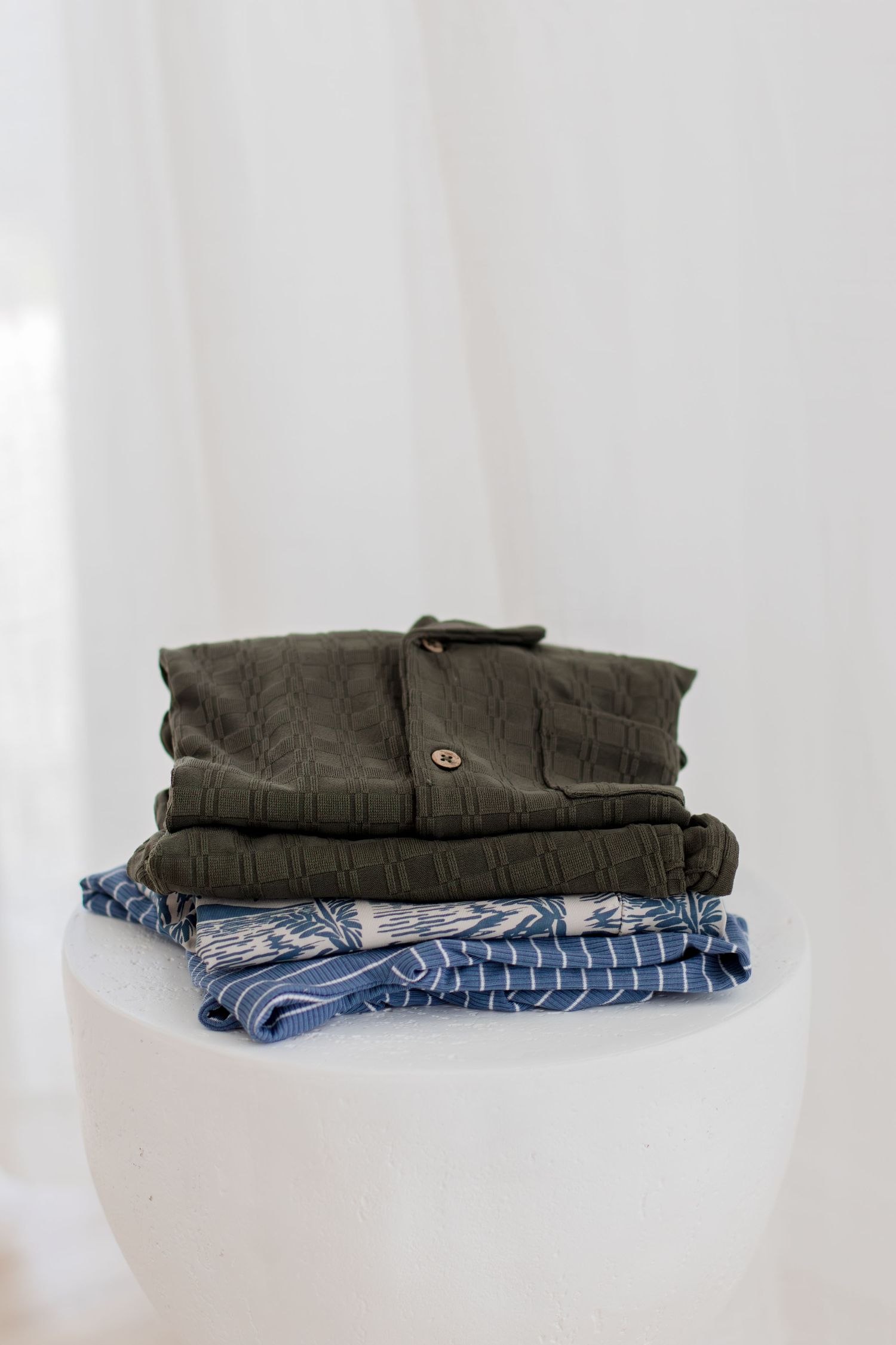 Neatly folded stack of olive green and blue patterned clothing on white round pedestal display.