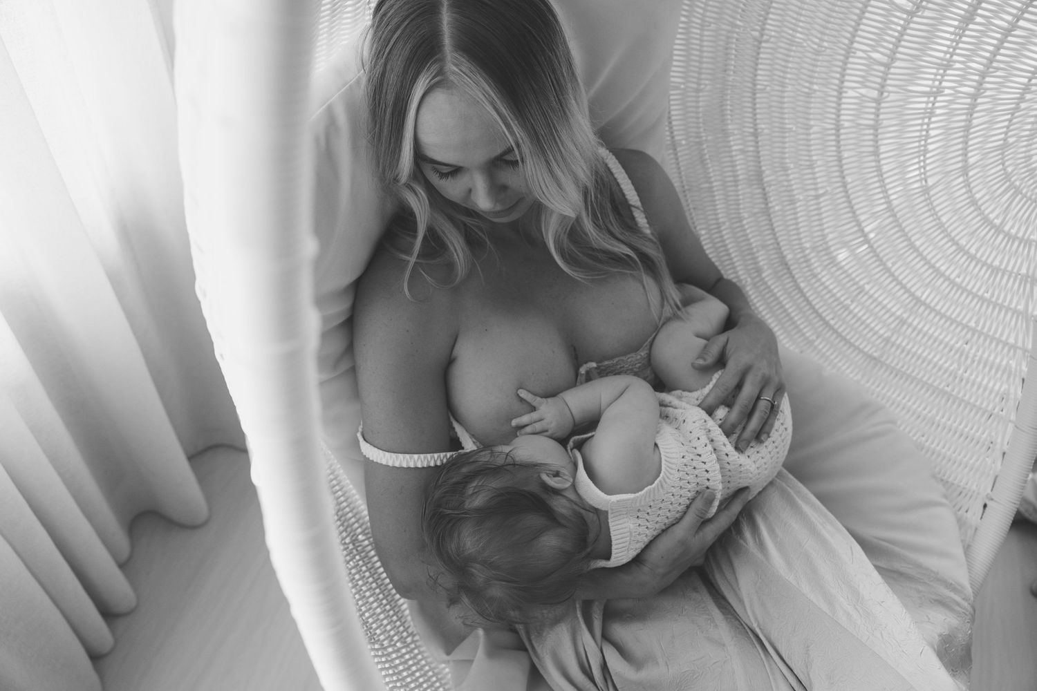 Black and white overhead photo of mother breastfeeding newborn baby in rattan chair.