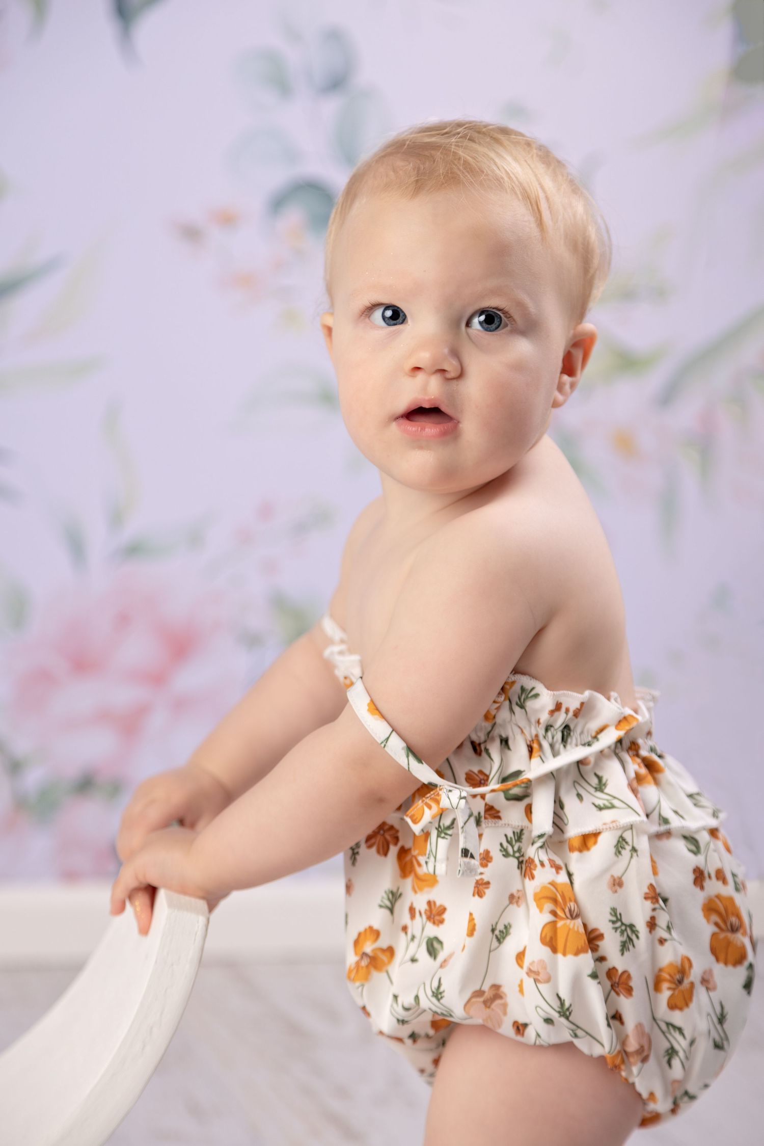 Adorable baby girl in floral bloomers standing against pastel floral wallpaper backdrop.