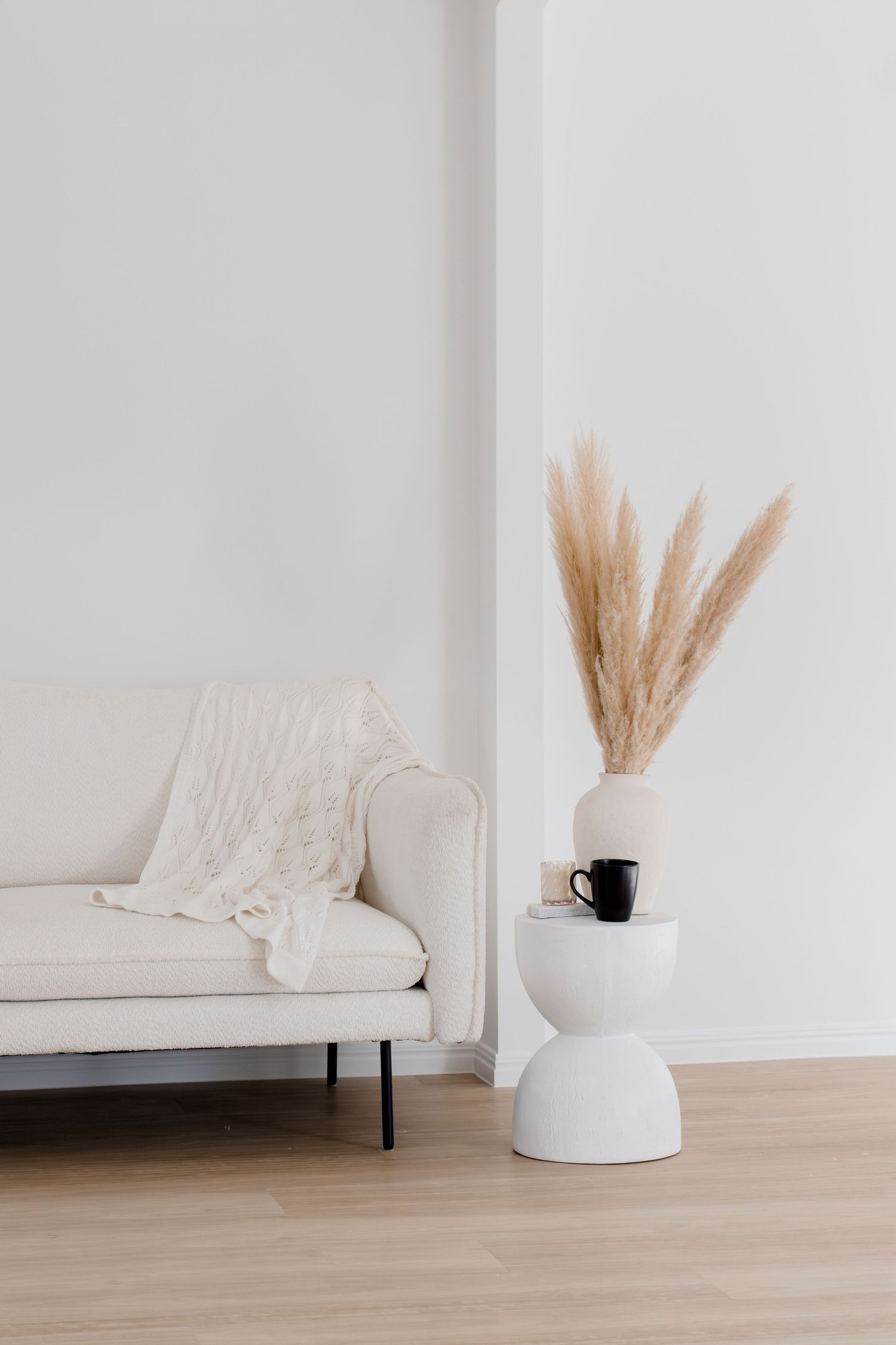 Cream sofa with blanket beside a white pedestal table holding pampas grass vase and black mug.