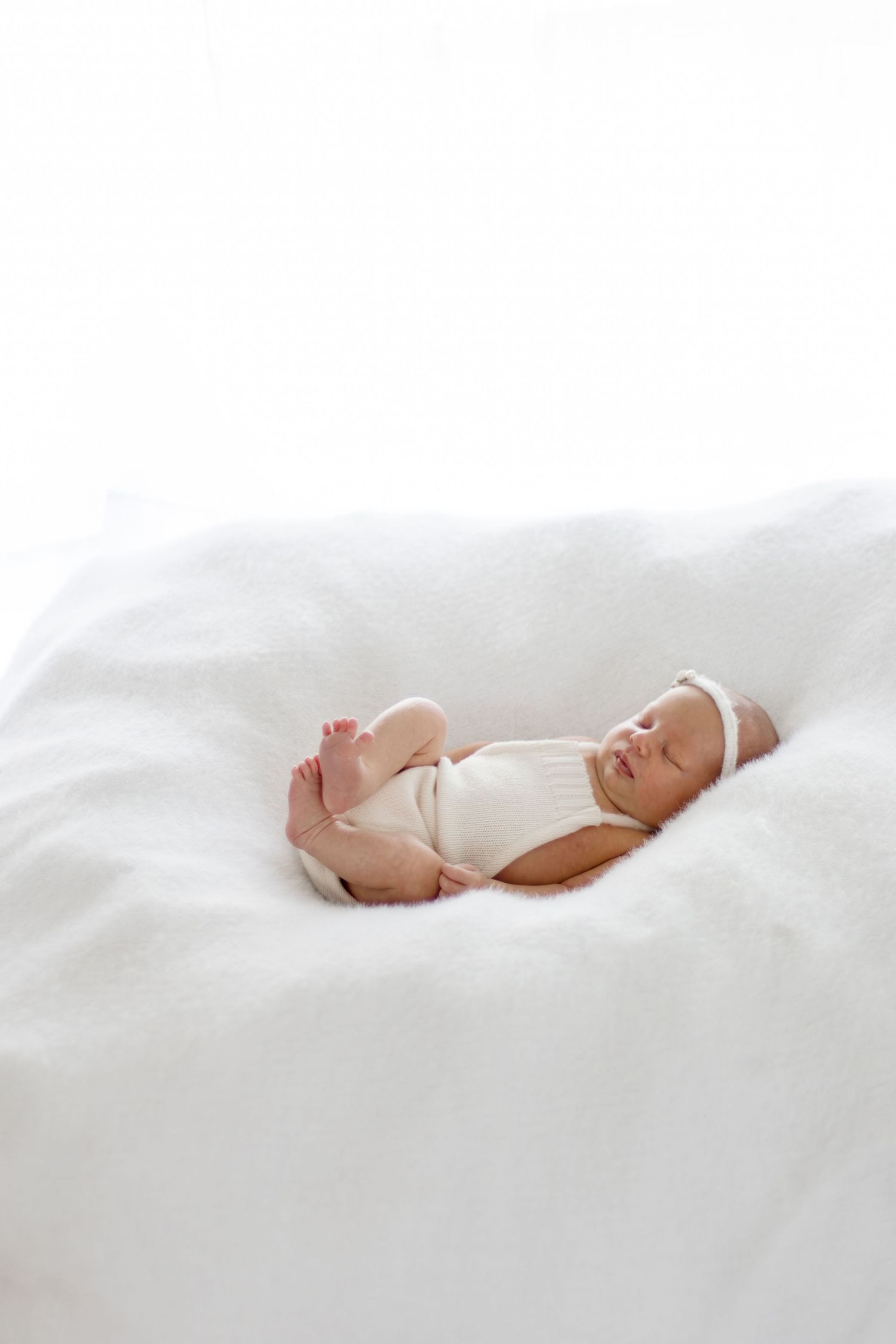 Peaceful newborn baby sleeping on white fluffy blanket wearing white onesie and headband.