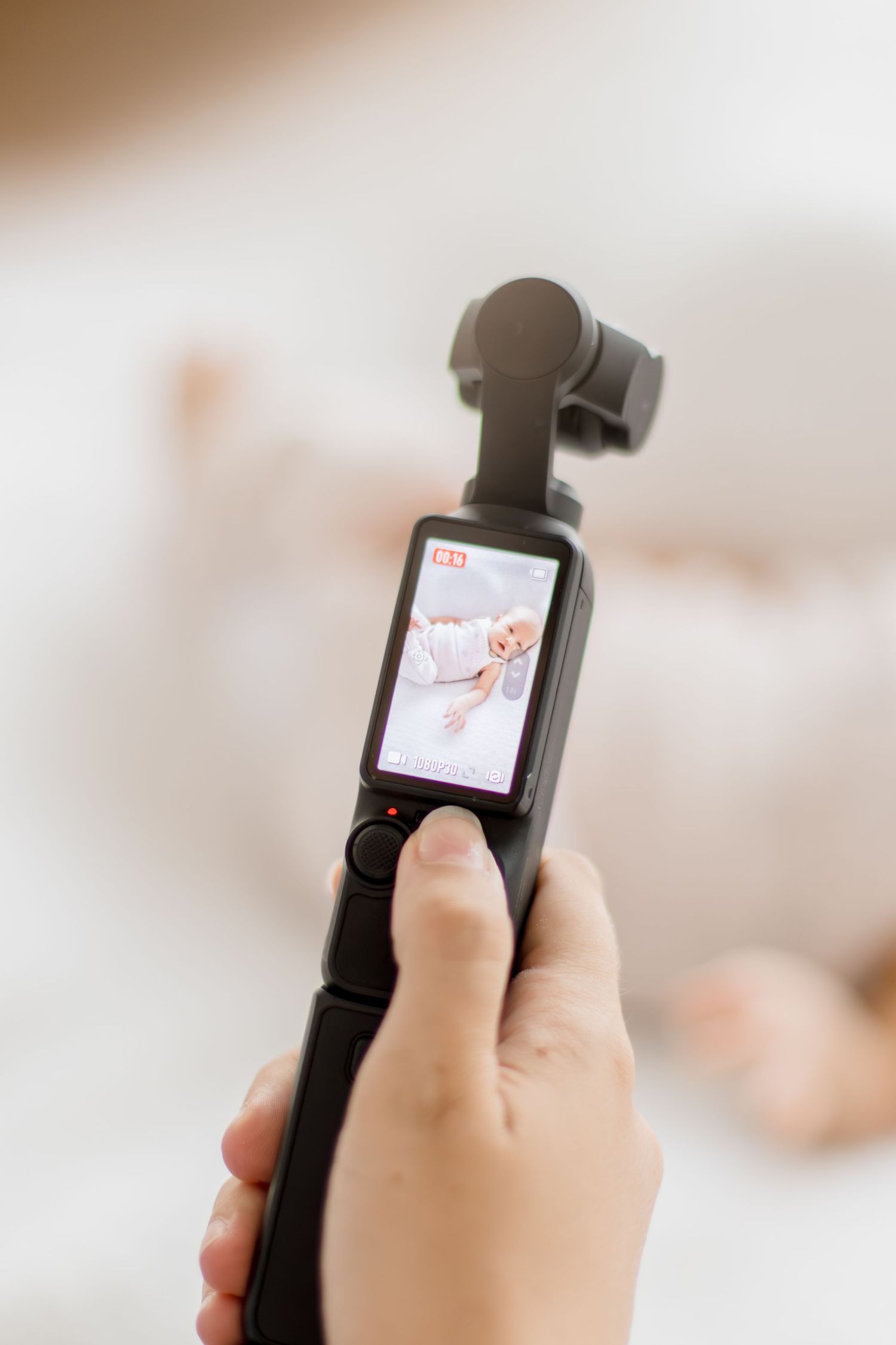 Hand holding a DJI Osmo Pocket camera filming a baby lying on a white bed, screen displaying live footage.