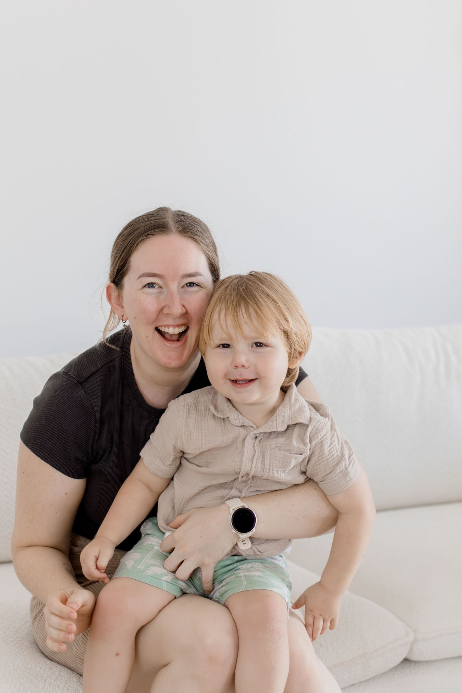 Happy mother and toddler boy sitting together on white couch smiling at camera in bright white room.