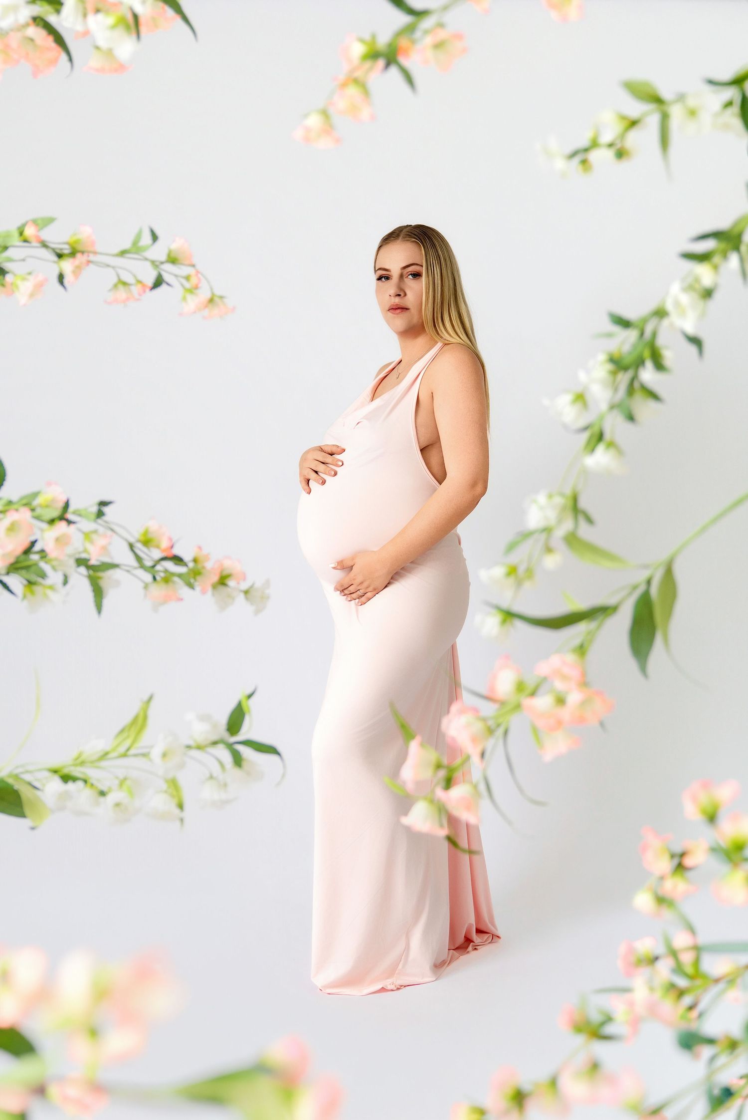 Pregnant woman in pink gown cradling belly, surrounded by delicate floral branches on white background.