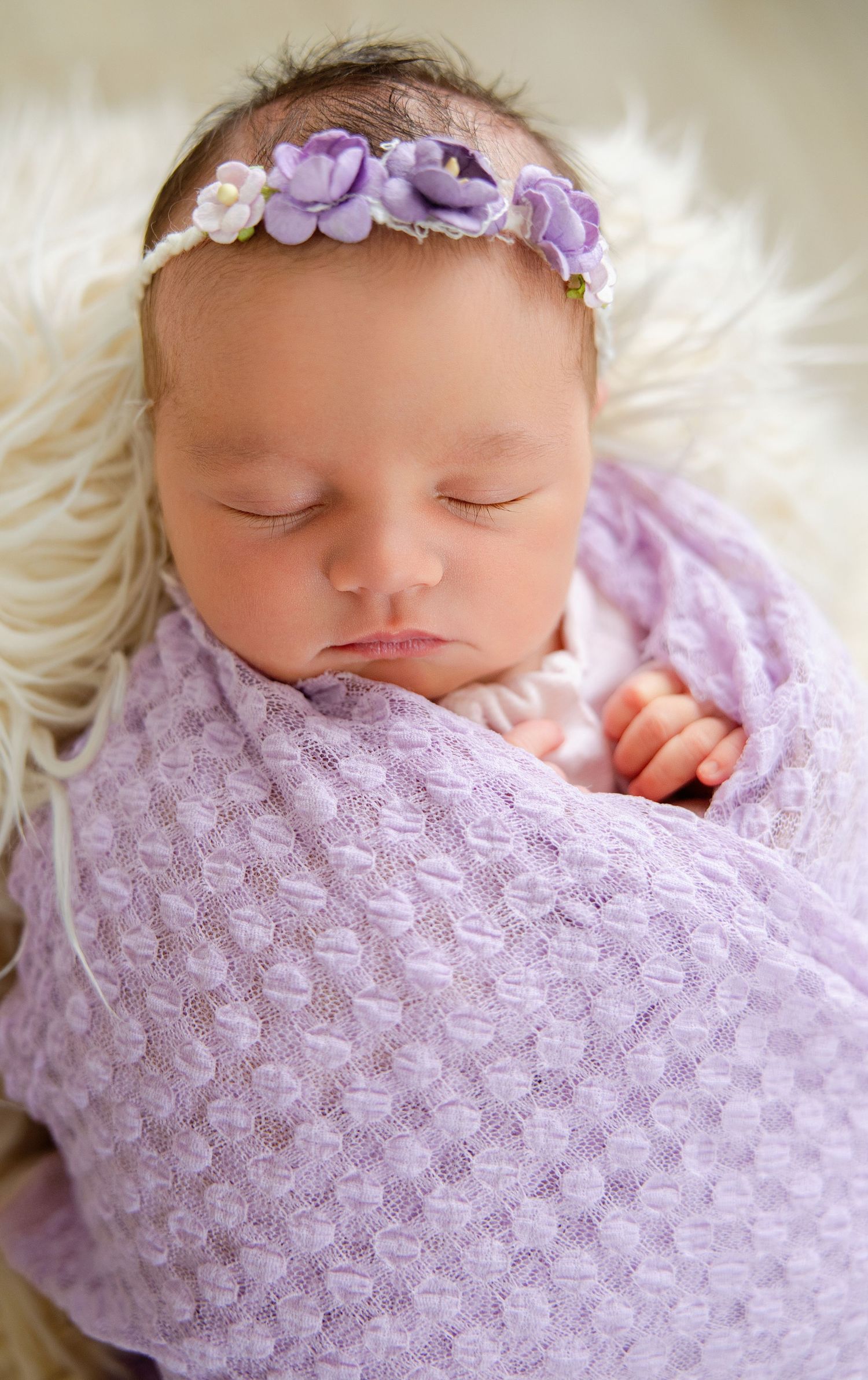 Sleeping newborn baby girl wrapped in lavender knit swaddle wearing purple floral headband on white fur backdrop.