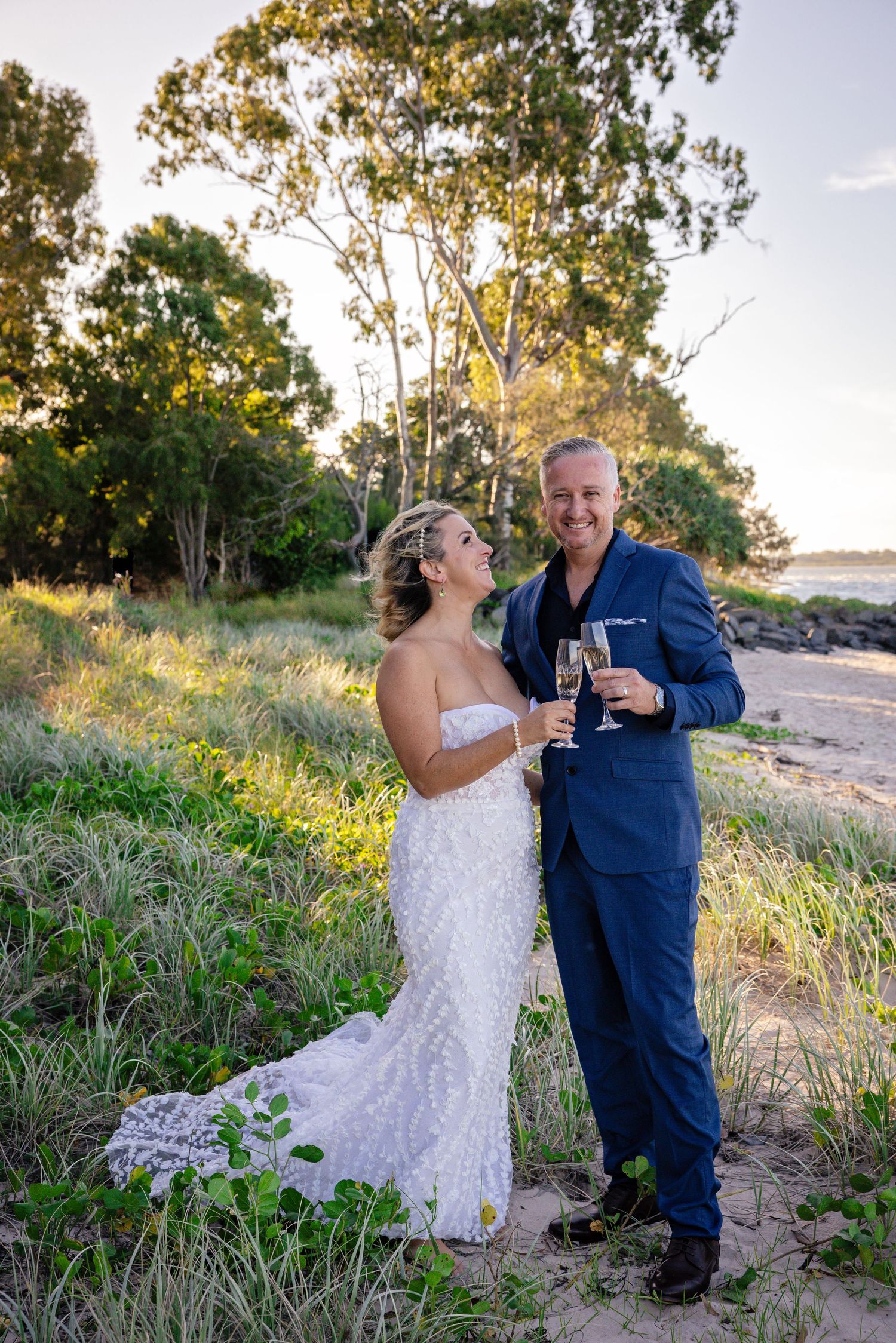 Bride and groom toast champagne at golden hour on scenic coastal beach setting.