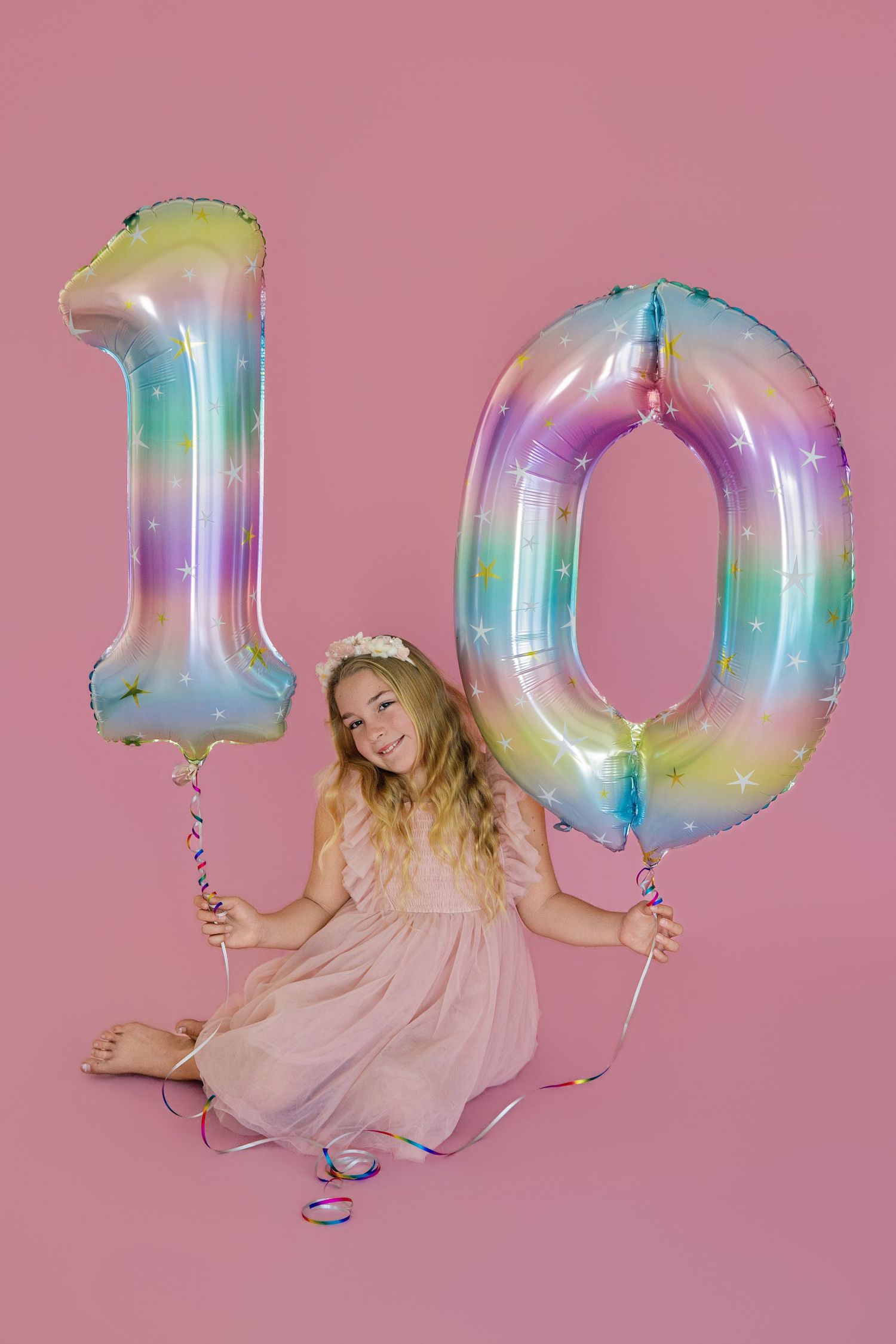 A smiling girl in a pink dress sits holding rainbow number 10 balloons on a pink background, celebrating her birthday.