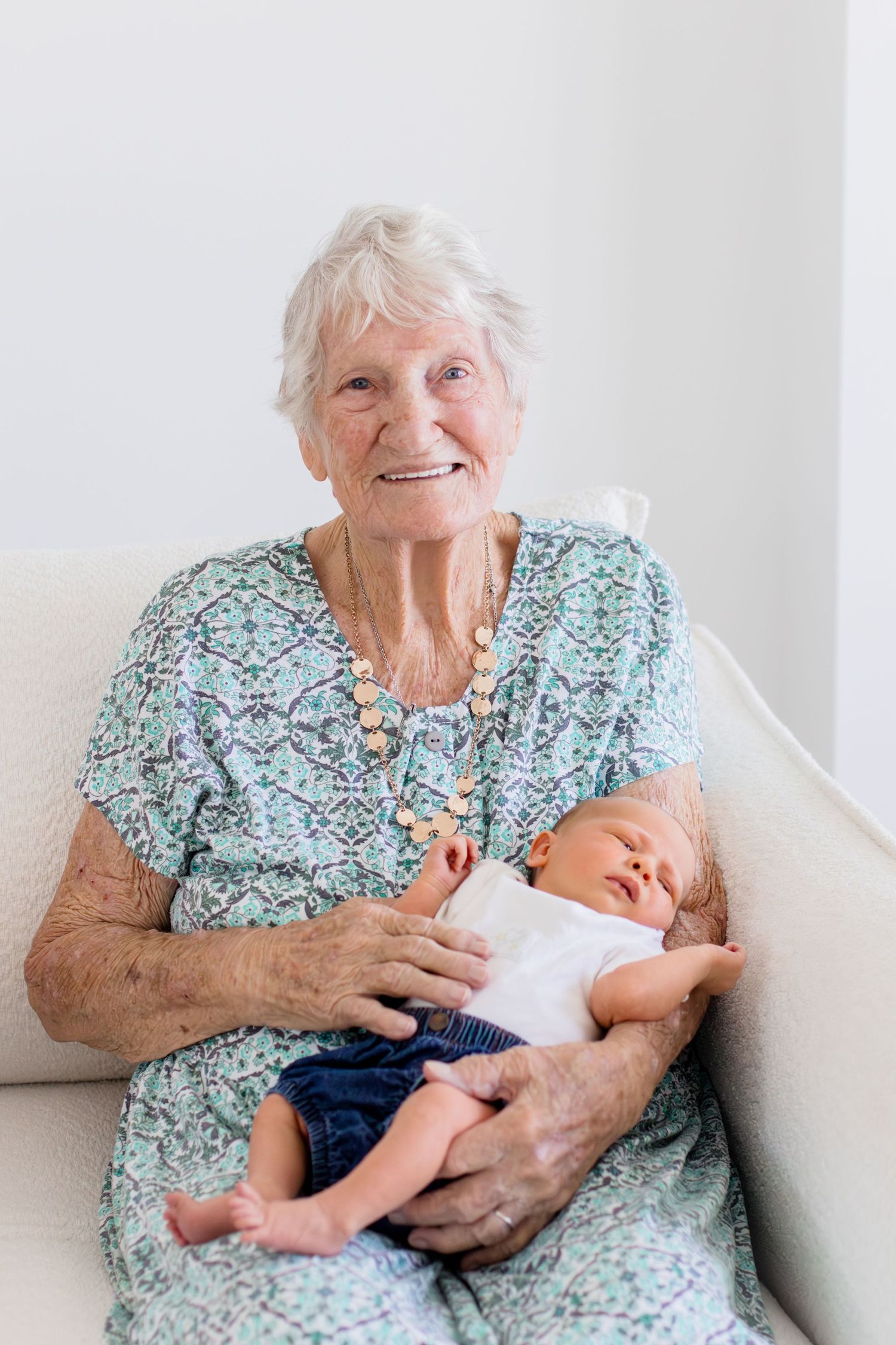Elderly grandmother smiling while holding sleeping newborn baby in white onesie on white armchair.