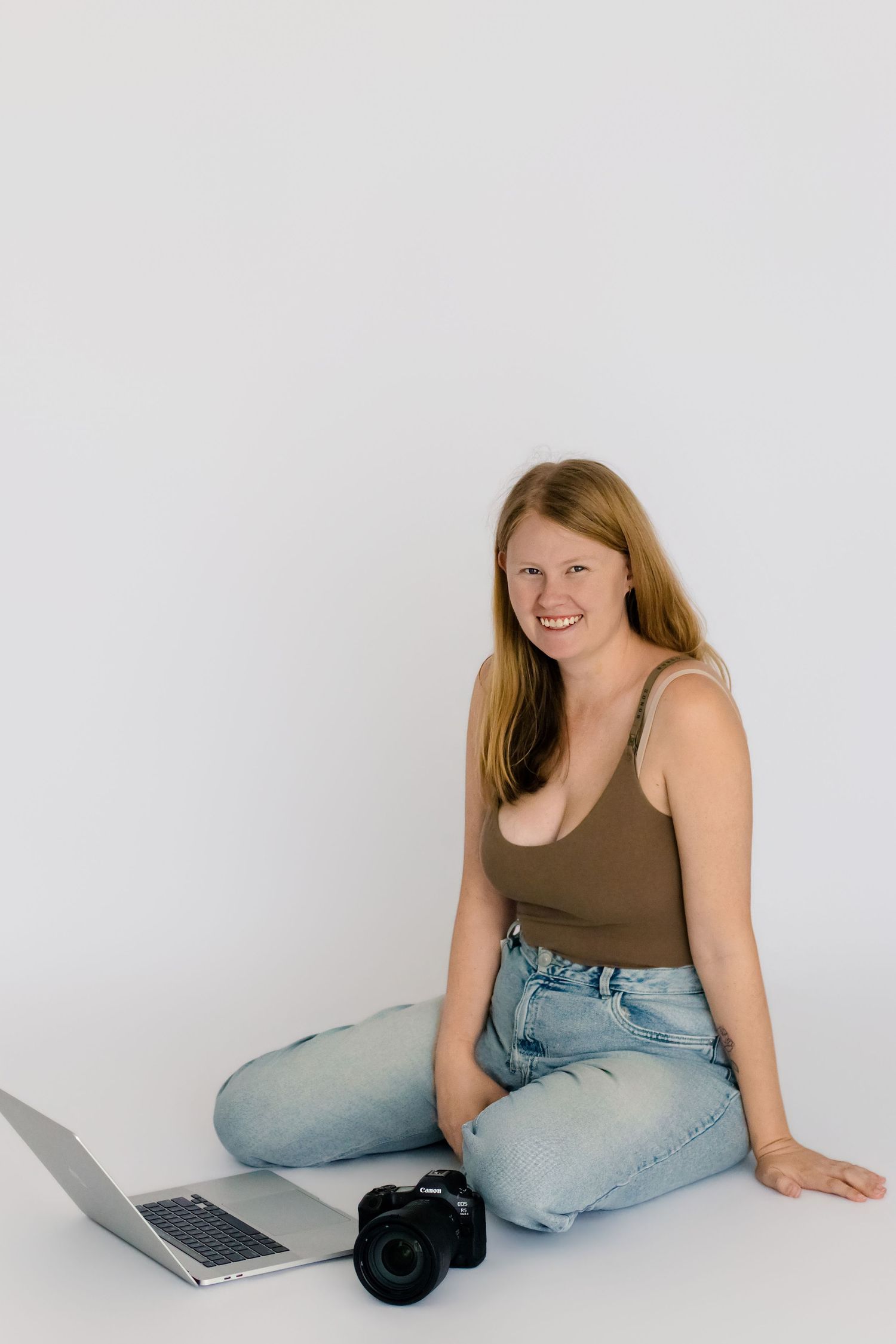 Smiling young woman sitting on white floor with laptop and camera, wearing brown tank top and light blue jeans.