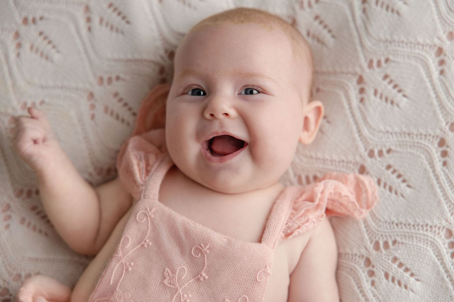 Smiling baby girl in pink dress lying on white knit blanket, happy infant with bright eyes and chubby cheeks.