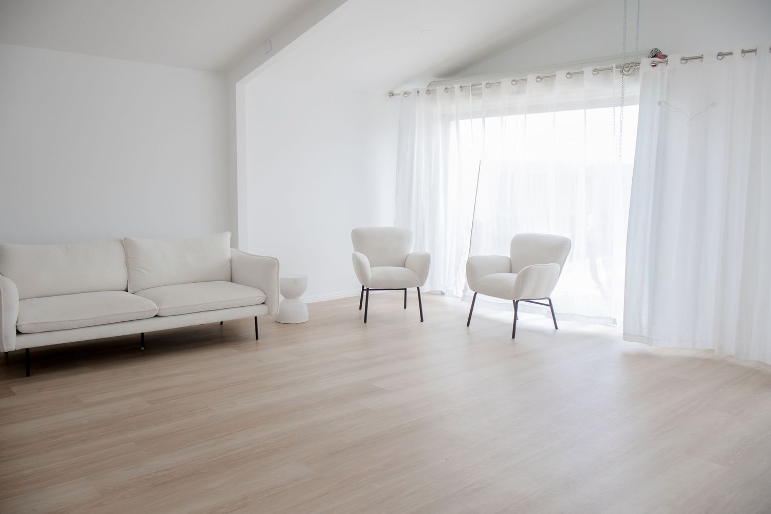 Bright white living room with a sofa, two armchairs, sheer curtains, and light hardwood flooring.