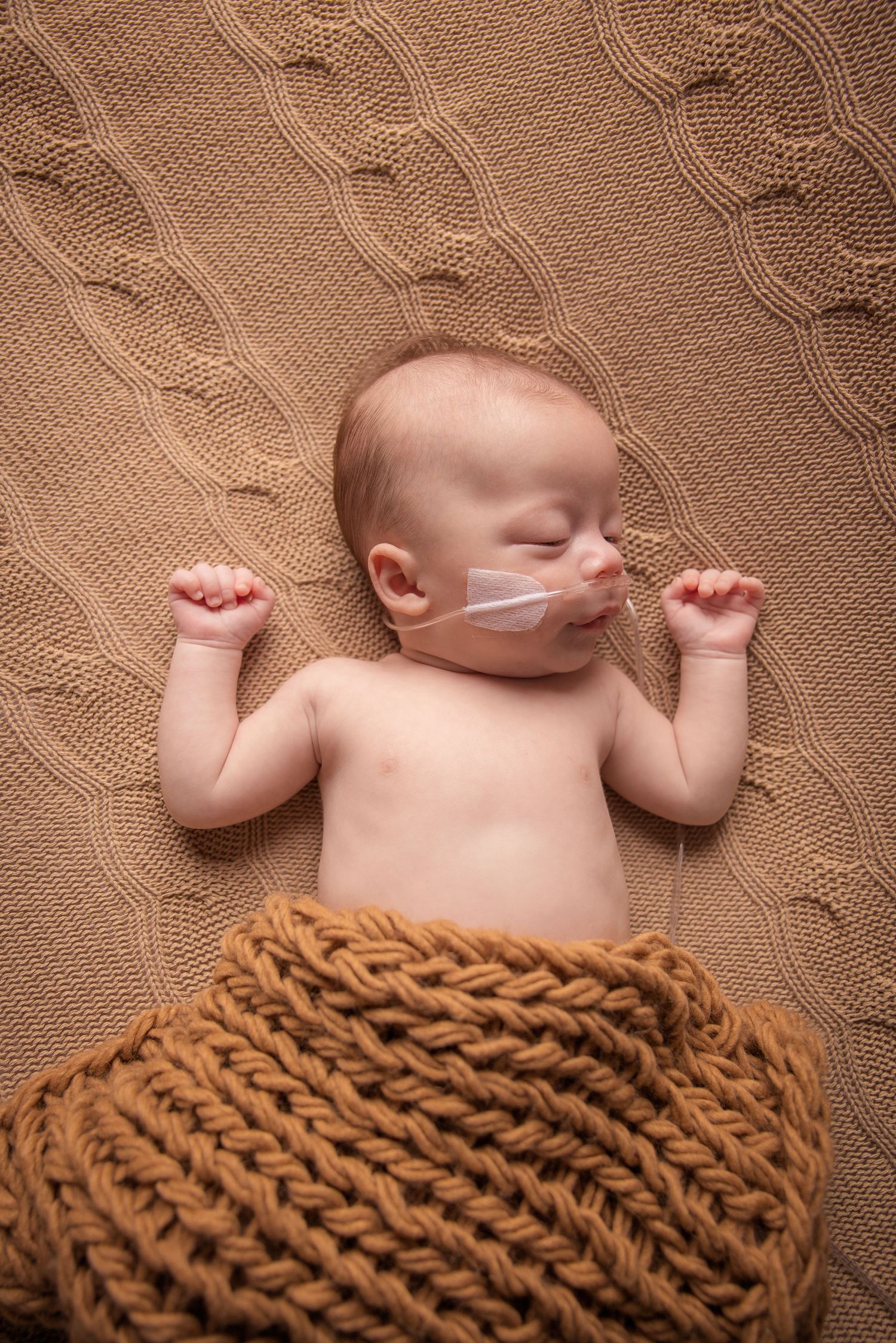 Sleeping newborn baby with nasal tube lying on brown knit blanket and wrap.