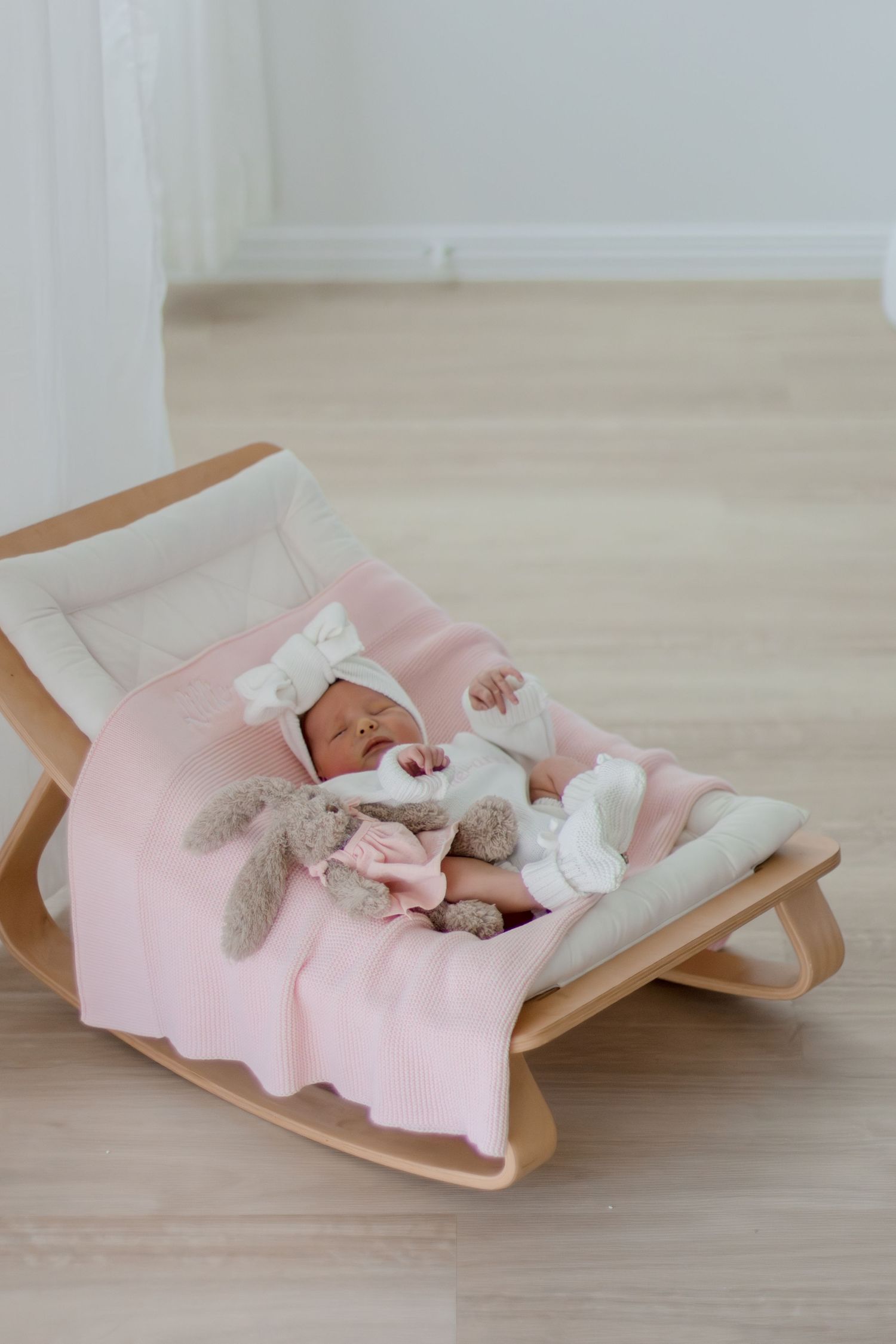 Sleeping newborn baby in white outfit with bow headband on pink blanket in wooden rocker with bunny toy.