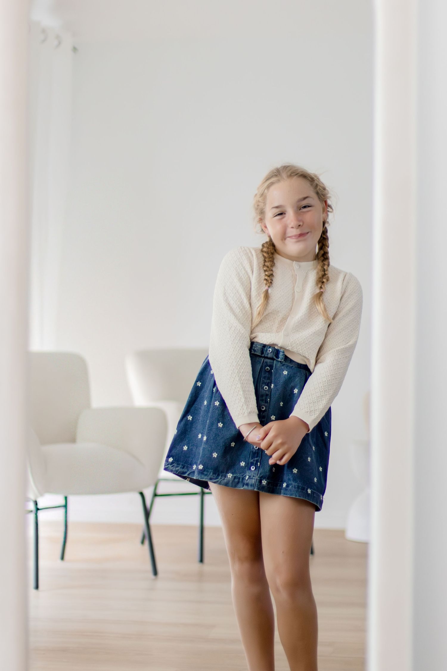 Smiling young girl with braids wearing cream sweater and denim skirt standing in bright white studio.