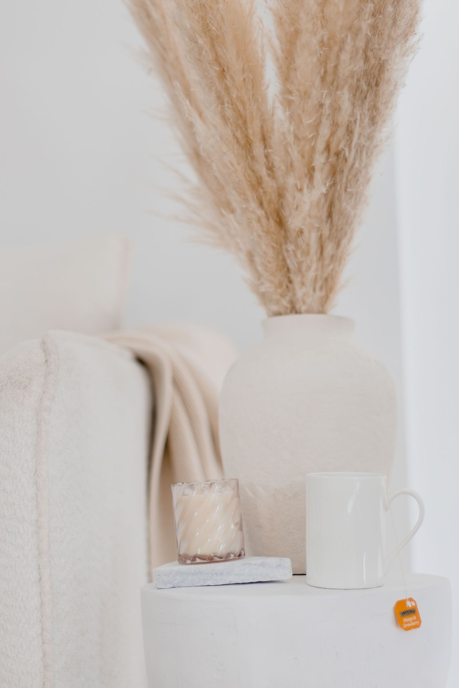 Close-up of beige pampas grass in ceramic vase beside a candle and white mug on pedestal.
