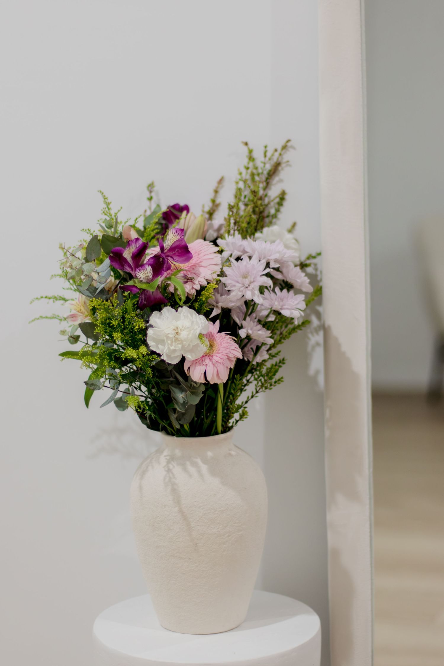 Elegant bouquet of purple, pink, and white flowers in a cream textured vase on white pedestal.