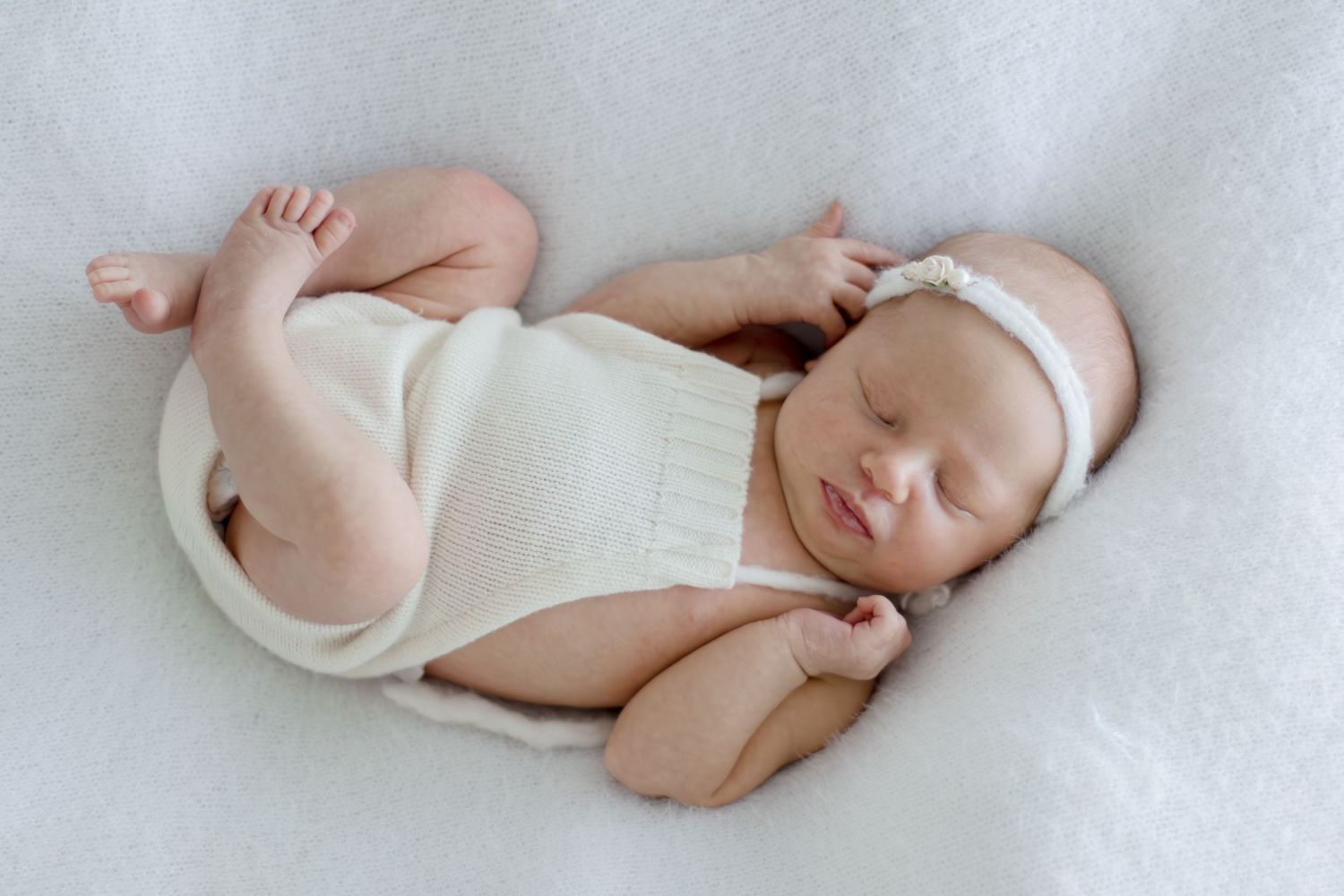 Peaceful newborn baby sleeping wrapped in cream knit, wearing white floral headband on soft white blanket.