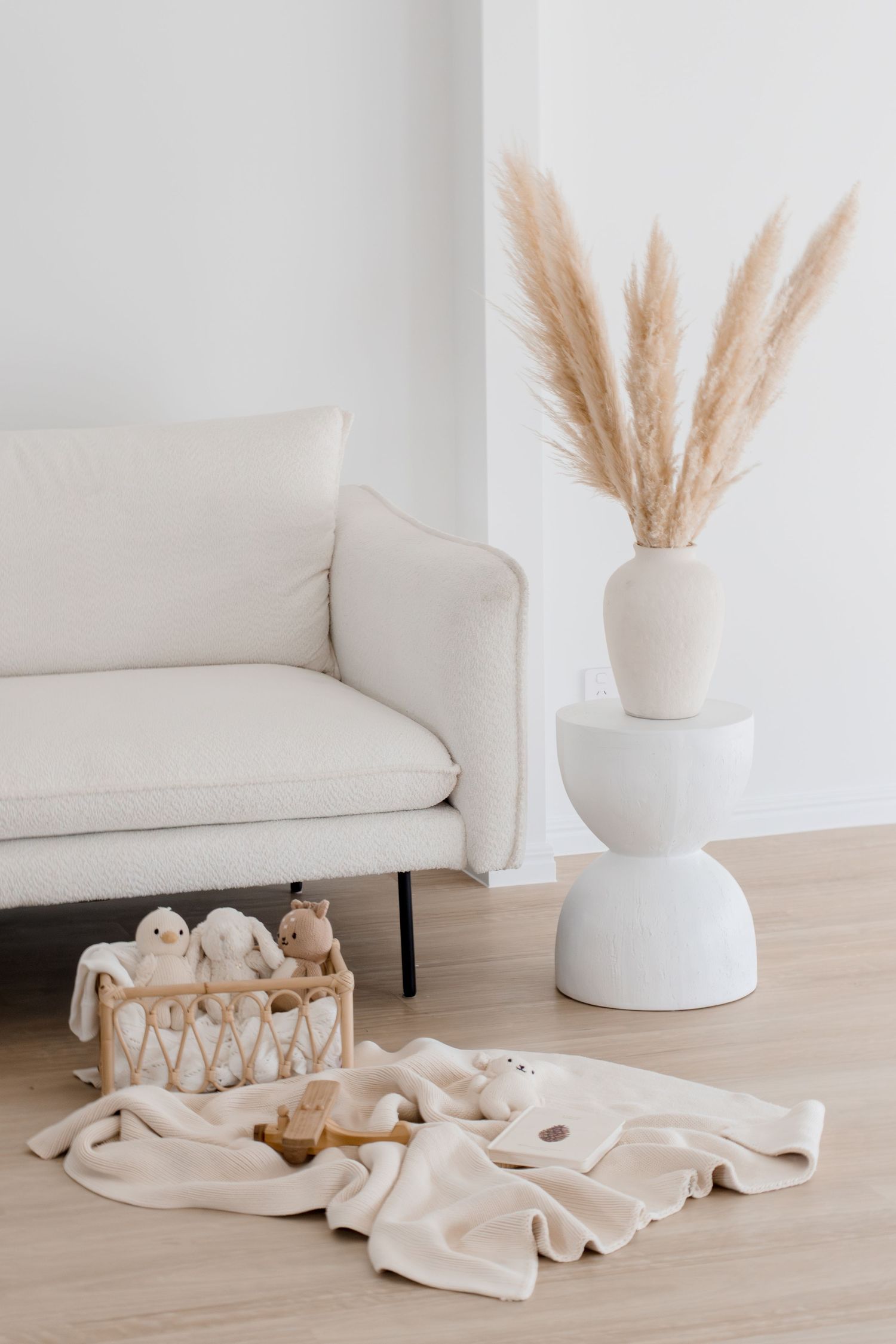 Minimalist nursery corner with white sofa, pampas grass, and neutral baby toys on floor.