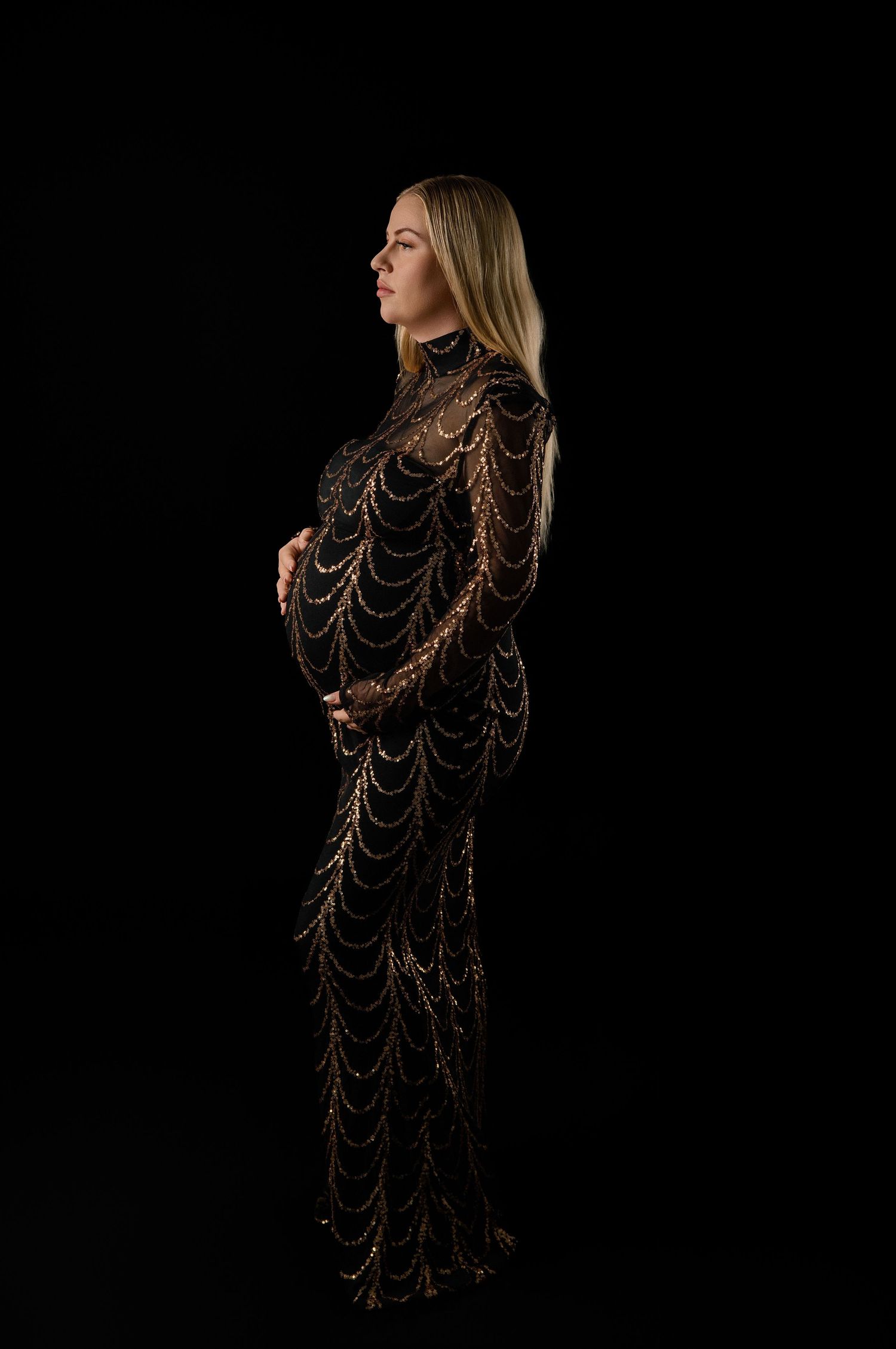 Pregnant woman in elegant black and gold beaded gown posing sideways against dark background.
