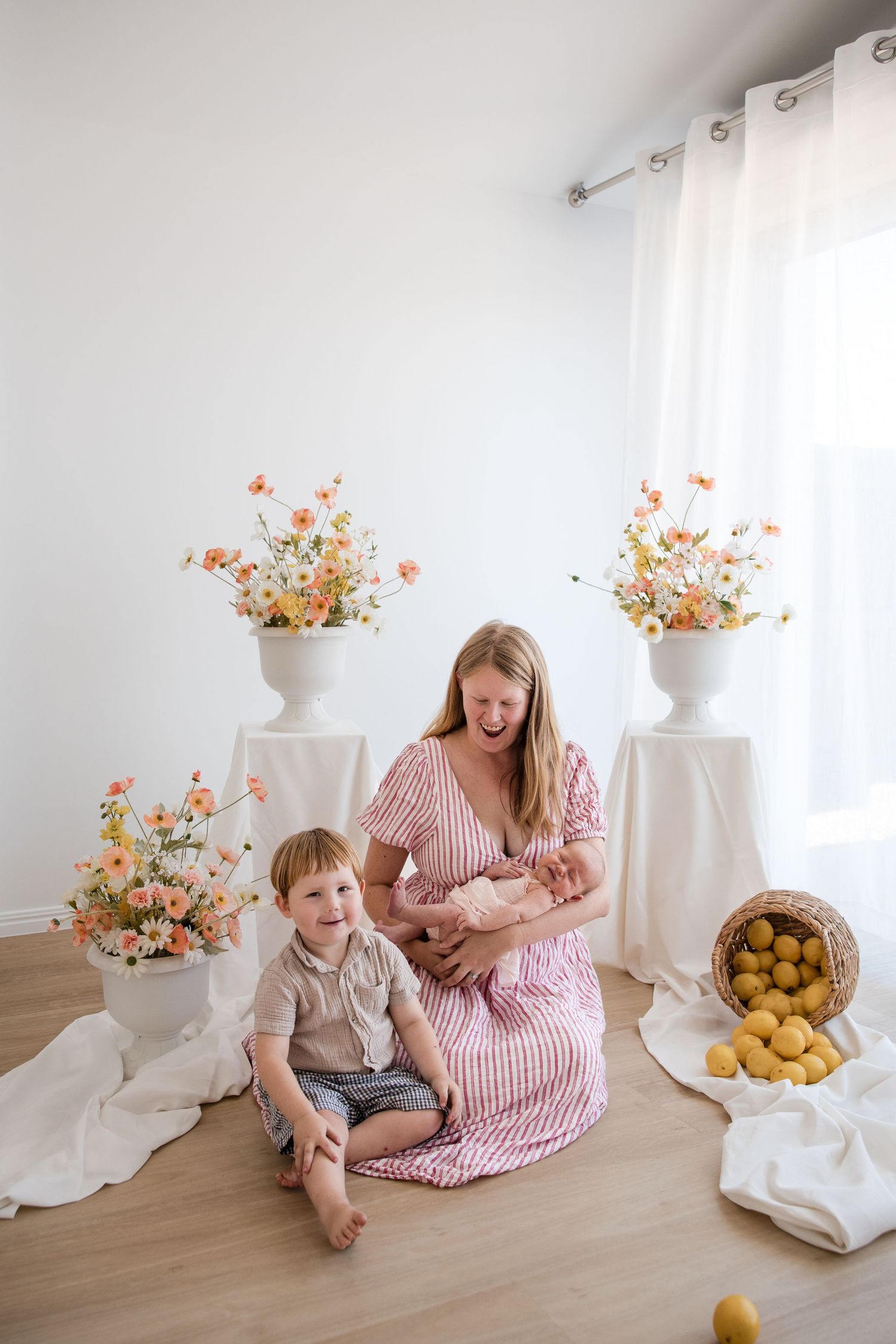 A mother in a pink striped dress holds her newborn while her toddler son sits beside her, surrounded by floral arrangements.