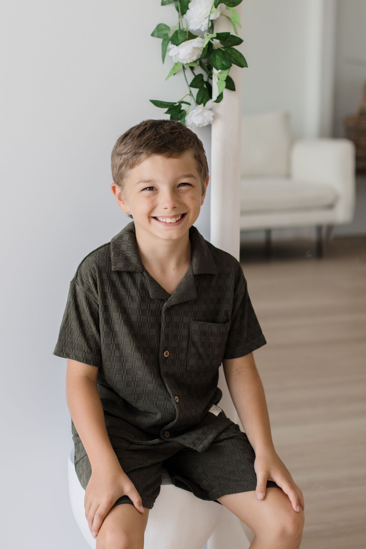 Smiling boy in dark green short sleeve button-up shirt and shorts sitting on white stool in bright studio.
