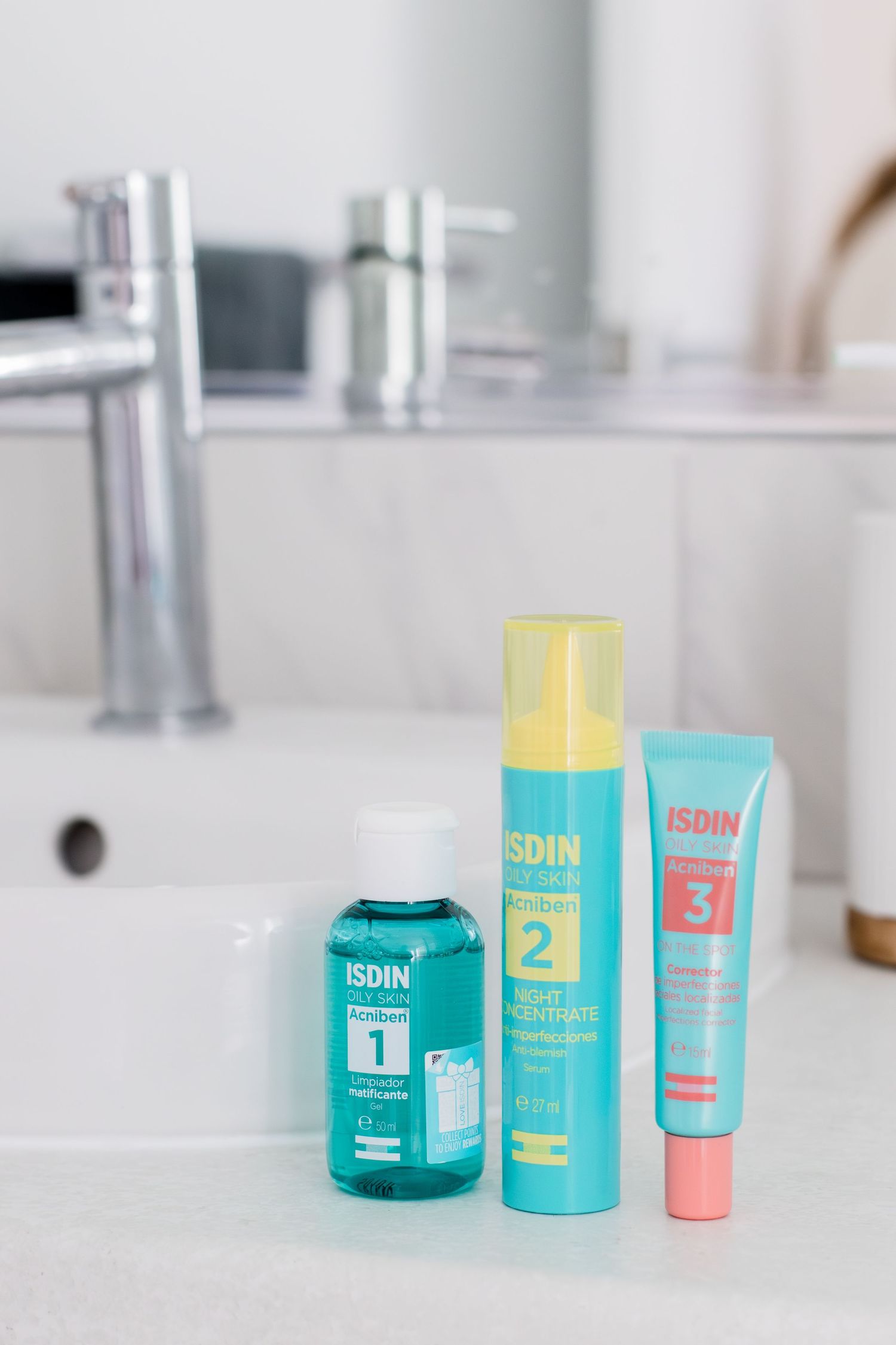 Three ISDIN skincare products in teal packaging displayed on white bathroom vanity counter.