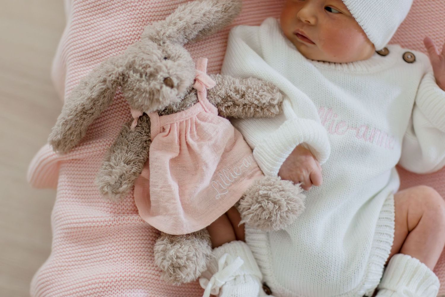 Close-up of newborn baby in white knit outfit beside fluffy gray bunny toy on pink blanket.