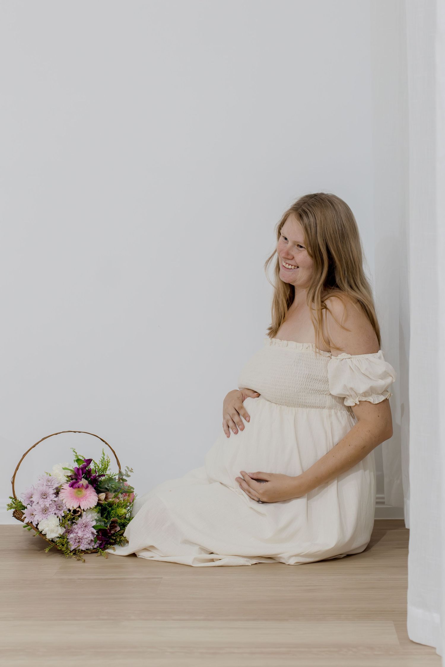 Smiling pregnant woman in white off-shoulder dress sitting on floor with floral bouquet beside her.