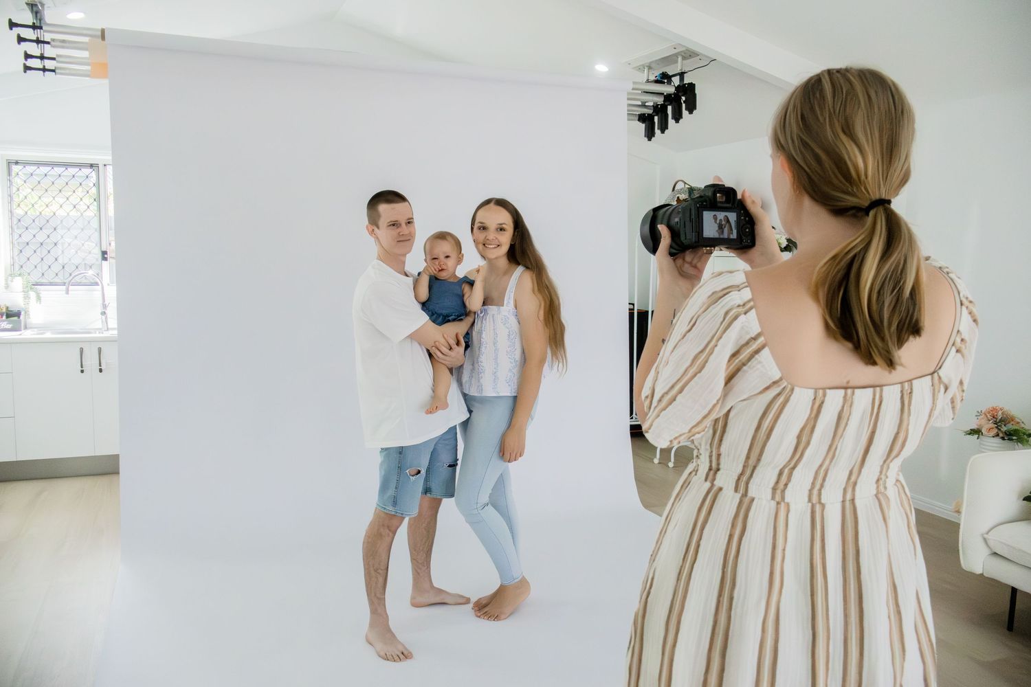 Photographer capturing family portrait of couple with baby in front of white backdrop in studio.