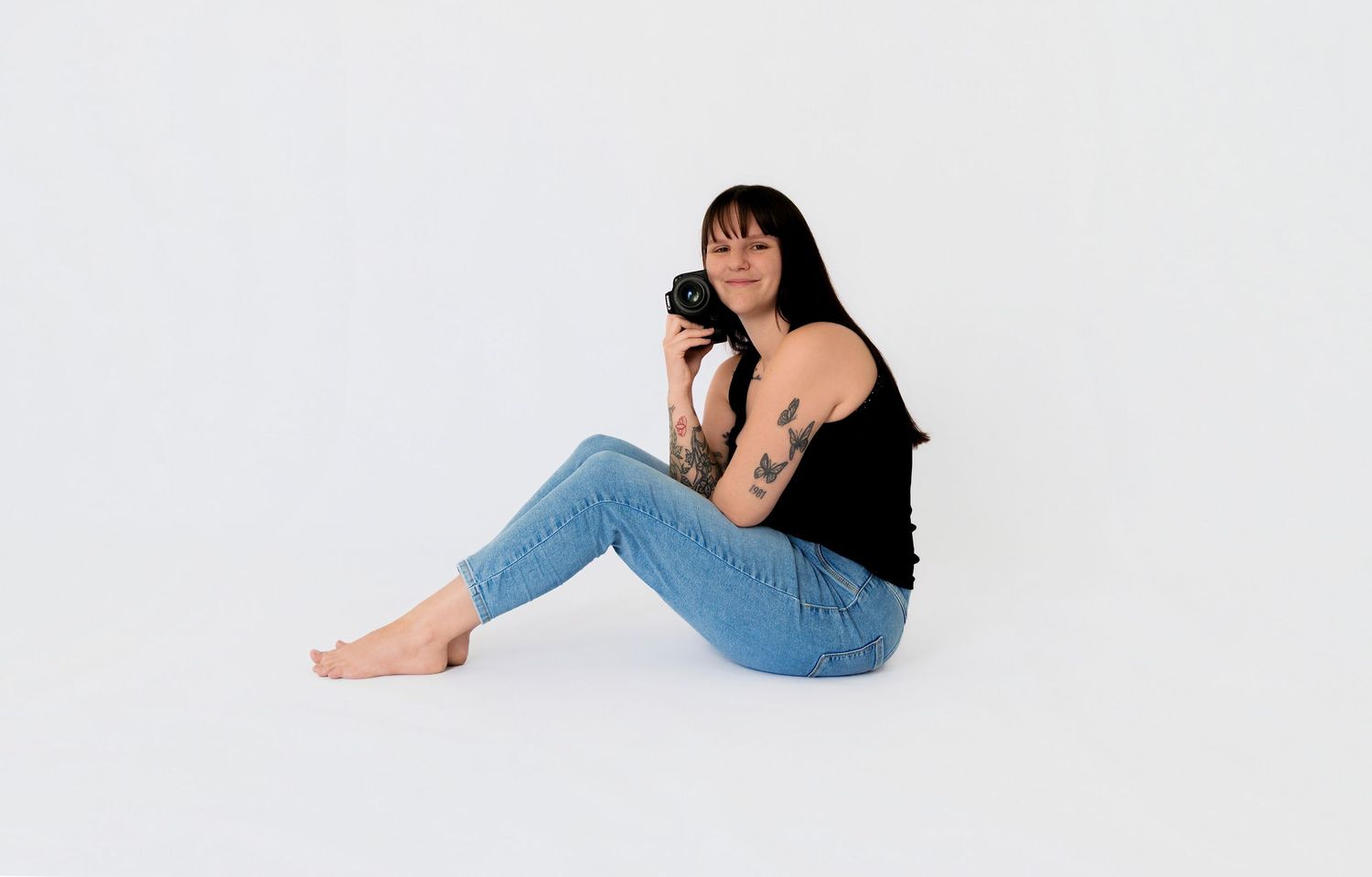 Woman with dark hair and tattoos sits on white floor holding a camera, wearing black top and jeans.