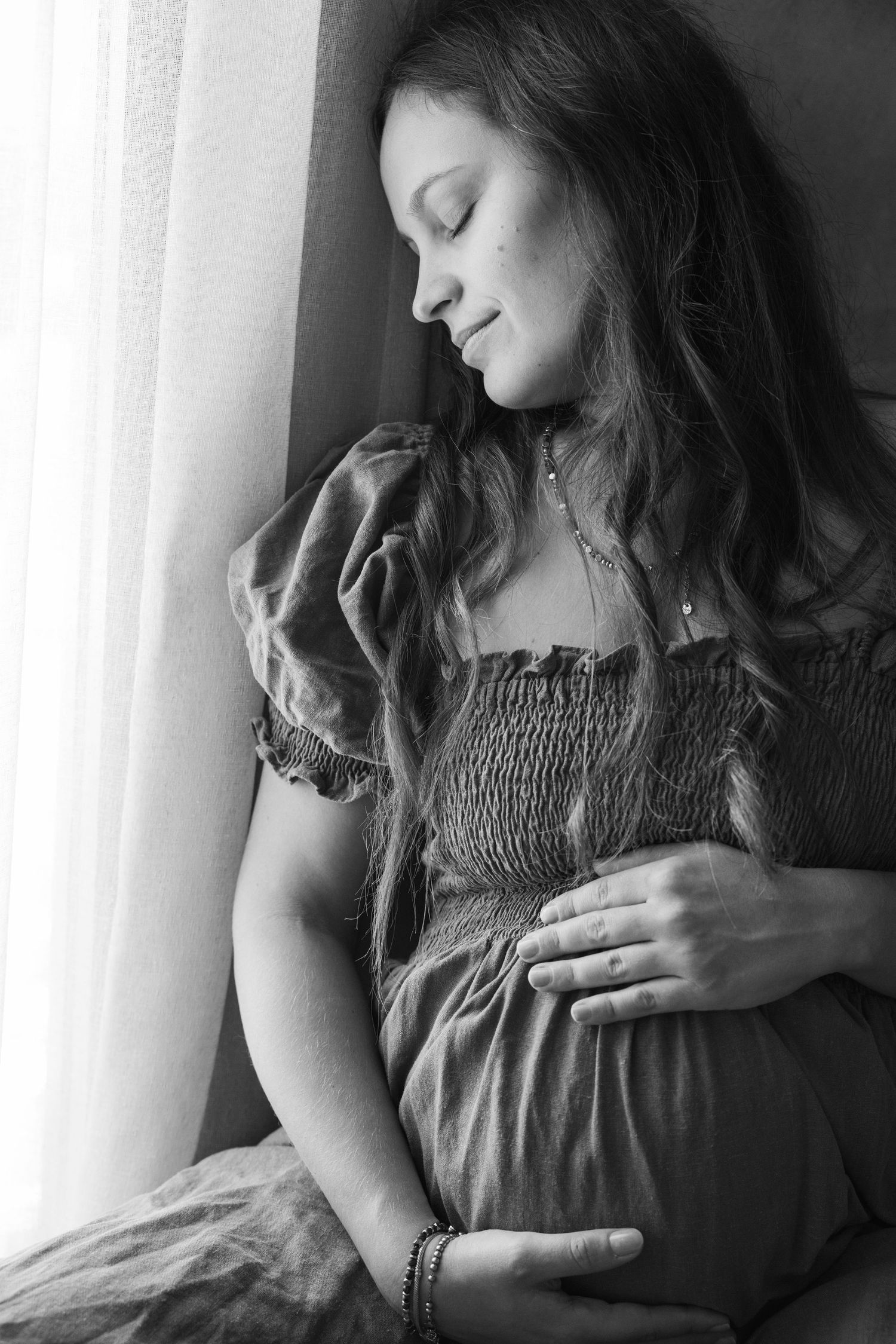 Black and white portrait of pregnant woman cradling baby bump by window in smocked dress.
