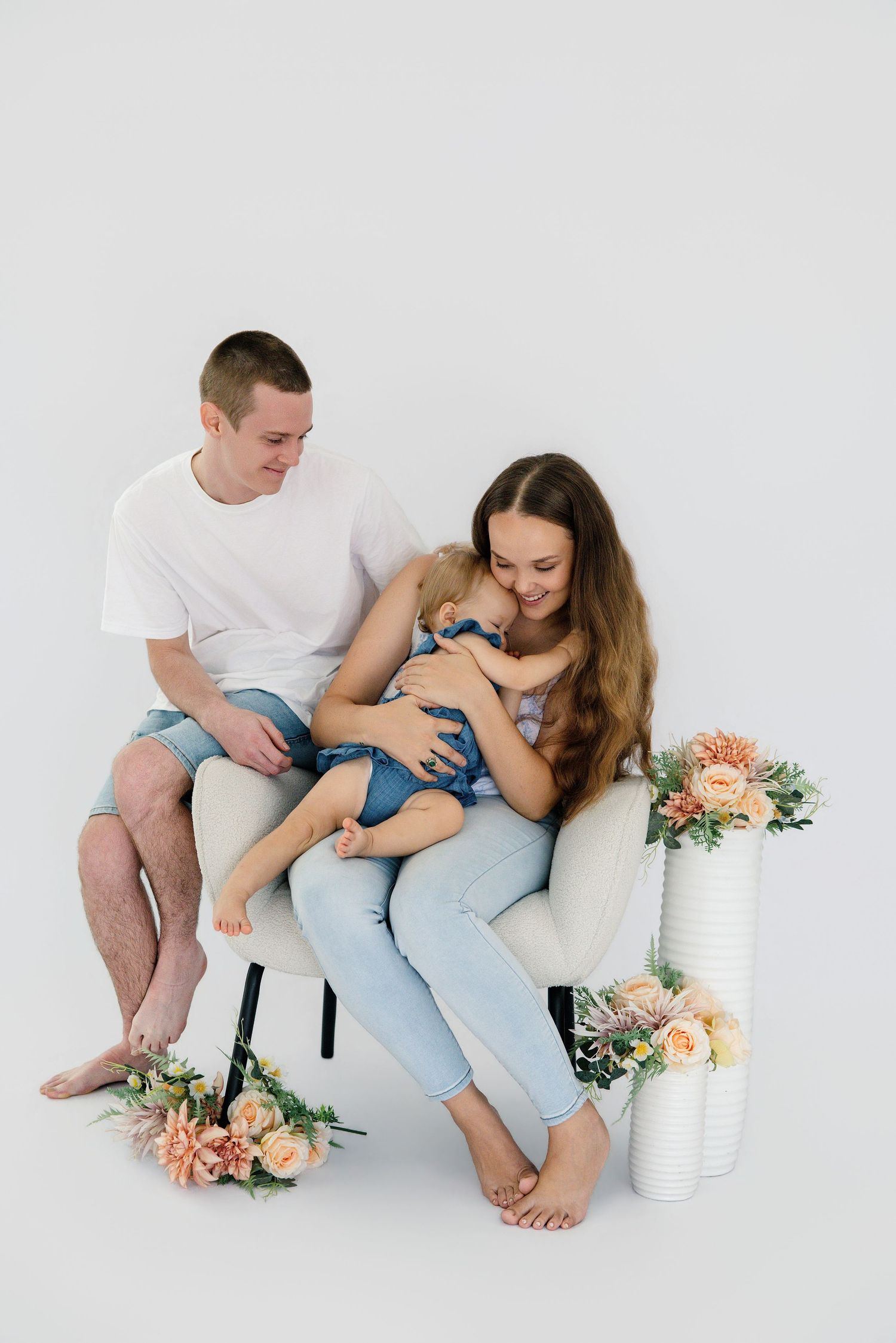 Young family with mother, father, and baby in denim sitting together in white armchair with flowers.