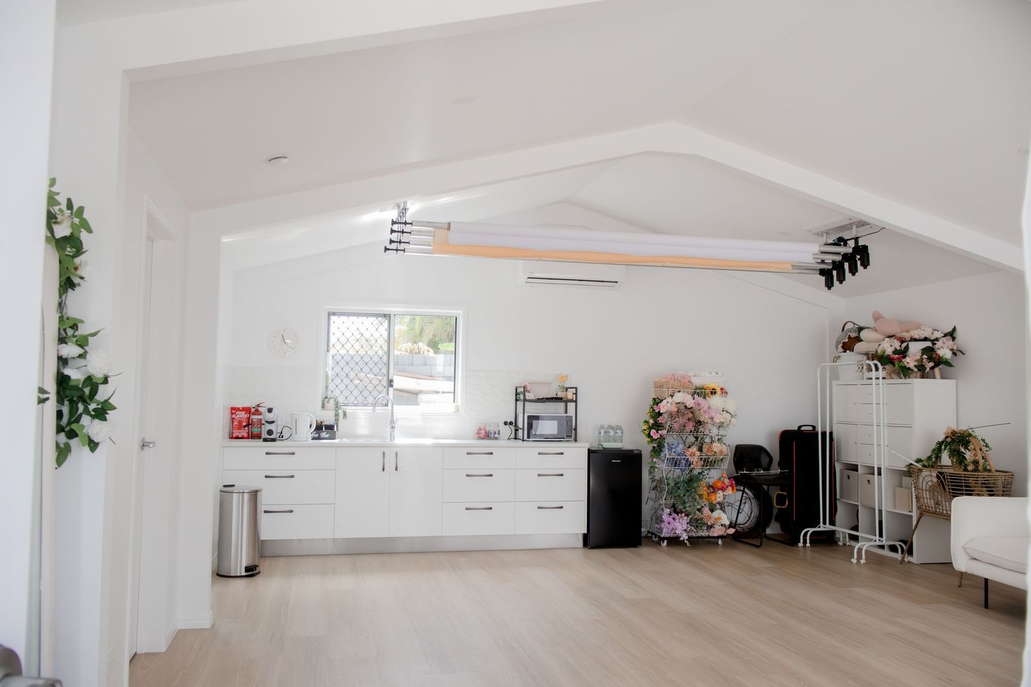 White studio space with vaulted ceiling, kitchen counter, lighting equipment, and floral decorations.