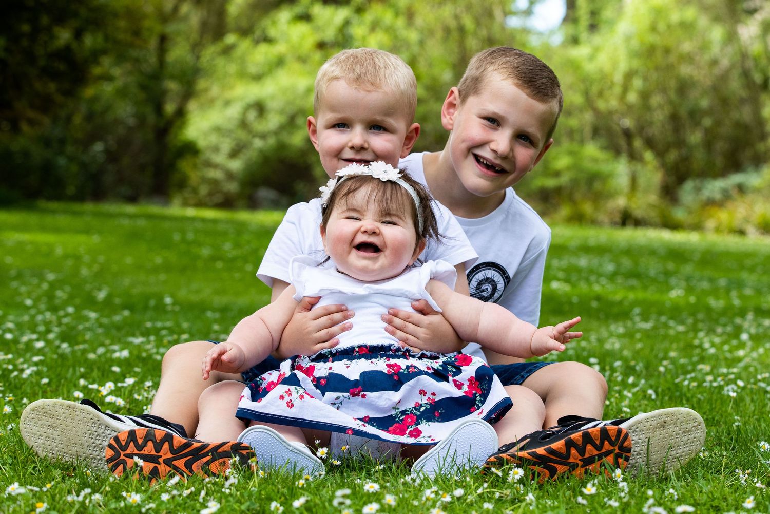 Three children sit together laughing on a grassy lawn surrounded by greenery on a sunny day.