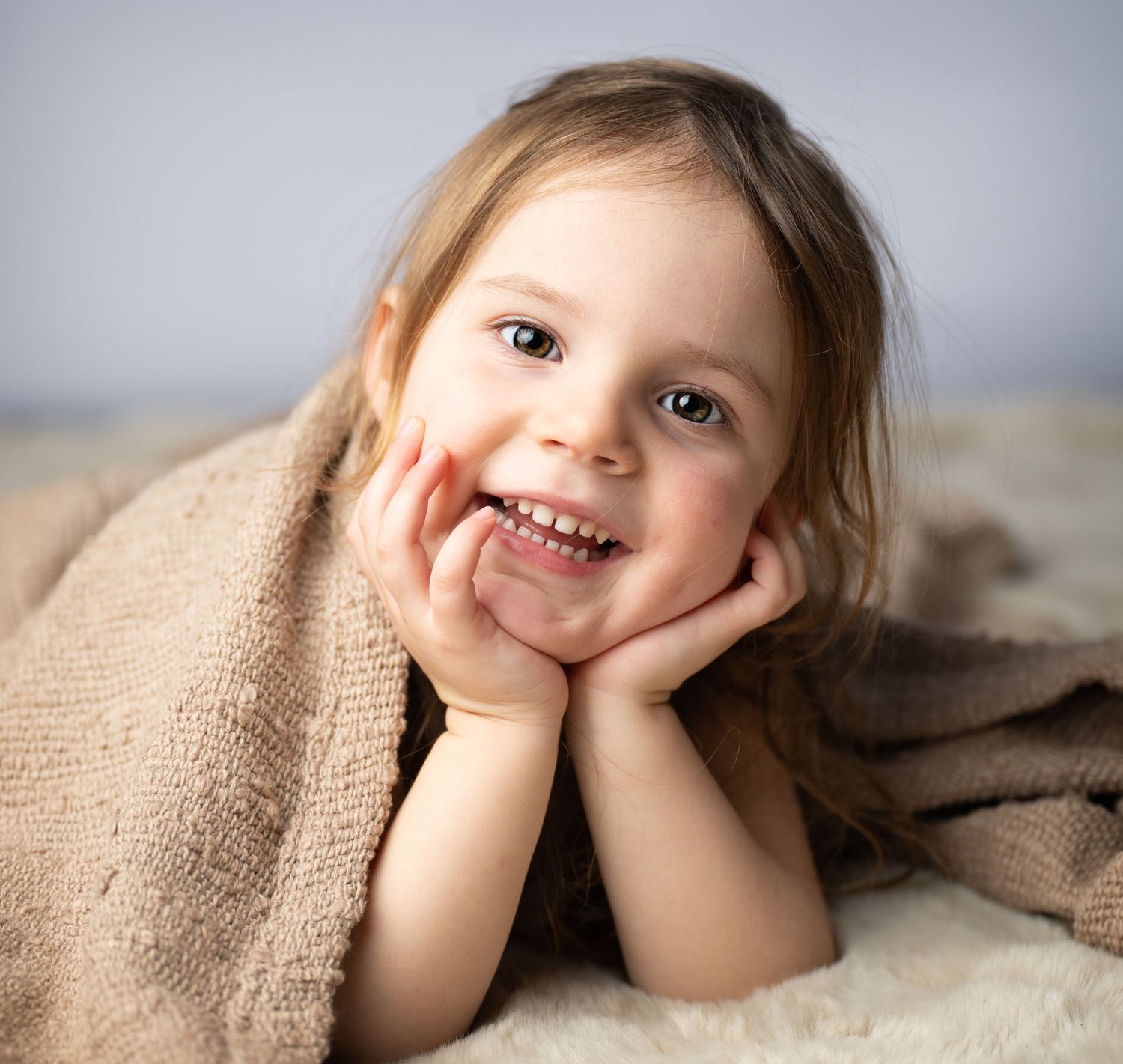 A cheerful young girl smiling with hands on cheeks, wrapped in a cozy brown blanket on a soft surface.