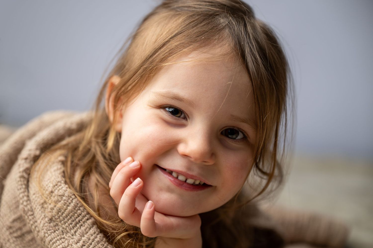 Smiling young girl with brown hair resting chin on hand, wearing a cozy beige sweater in a portrait photo.