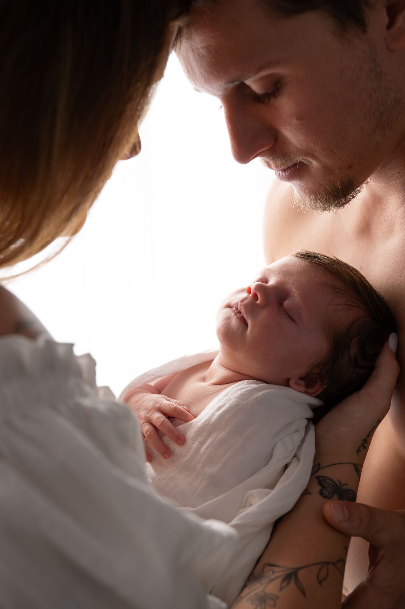 Mother and father gazing at newborn held between them in tender moment natural light in home session — Photos by Bailey Rose Gold Coast
