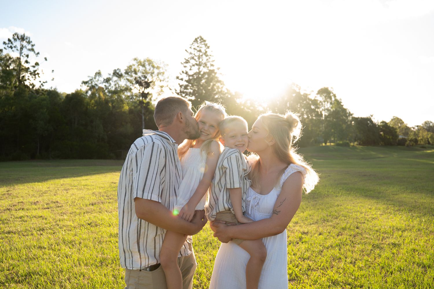 Family of four, children laughing in the middle of parents kissing their cheeks golden hour open field - Photos by Bailey Rose