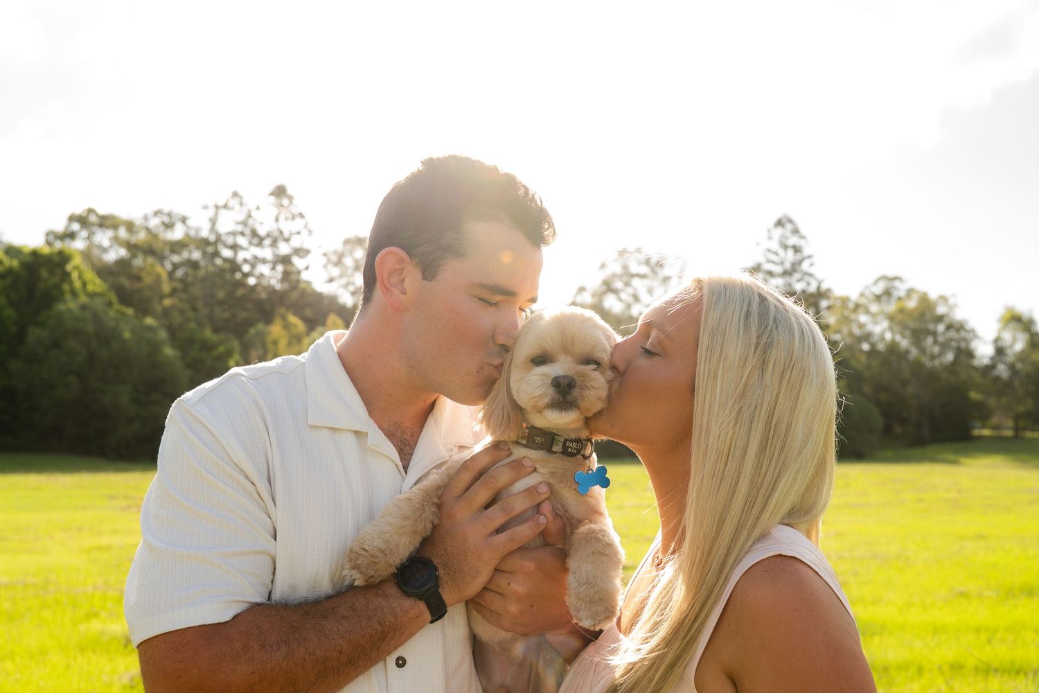 Couple kissing puppy during golden hour in an open field, unposed raw moments - Photos by Bailey Rose Brisbane
