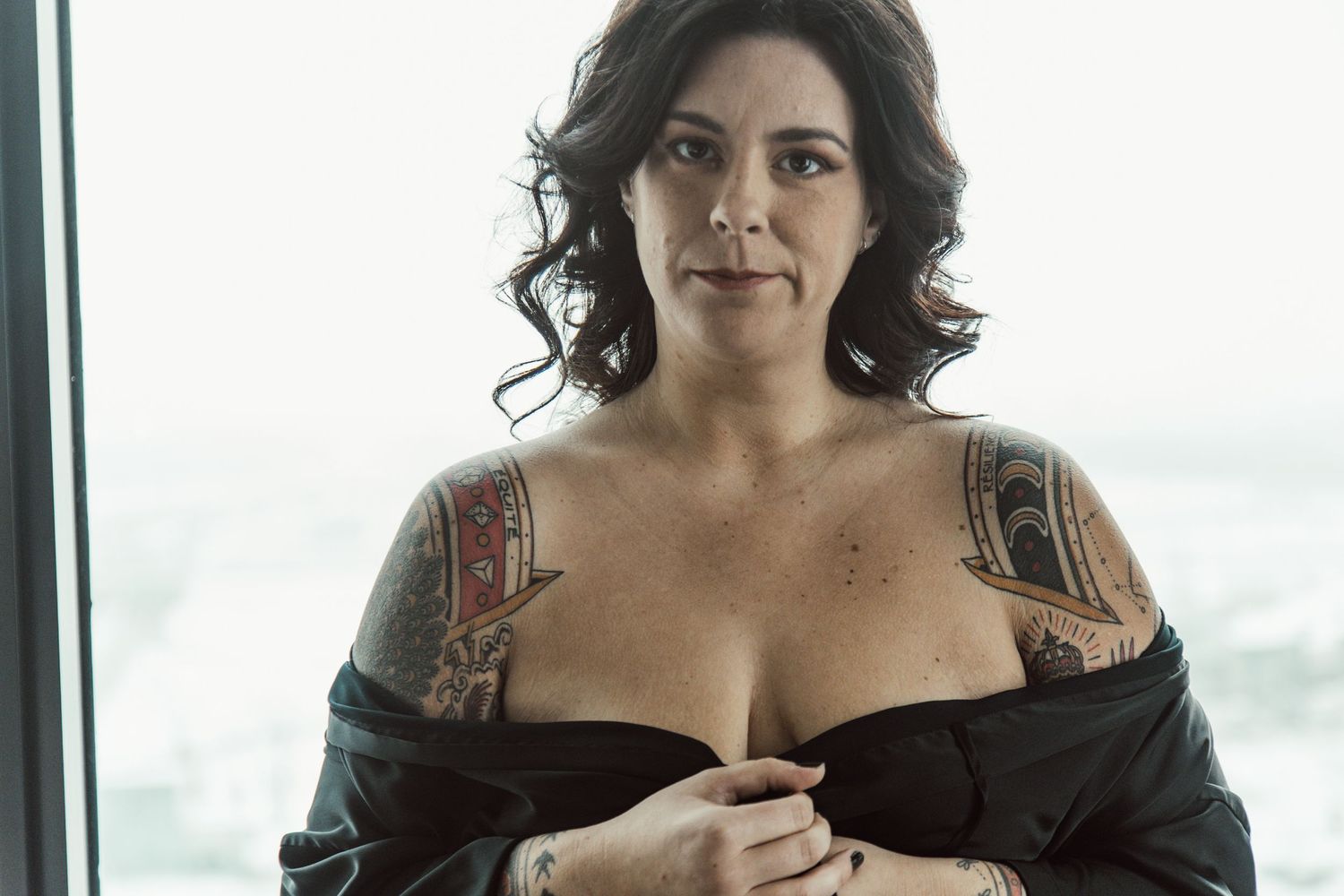 Artistic portrait by window showing shoulder tattoos and dark clothing.